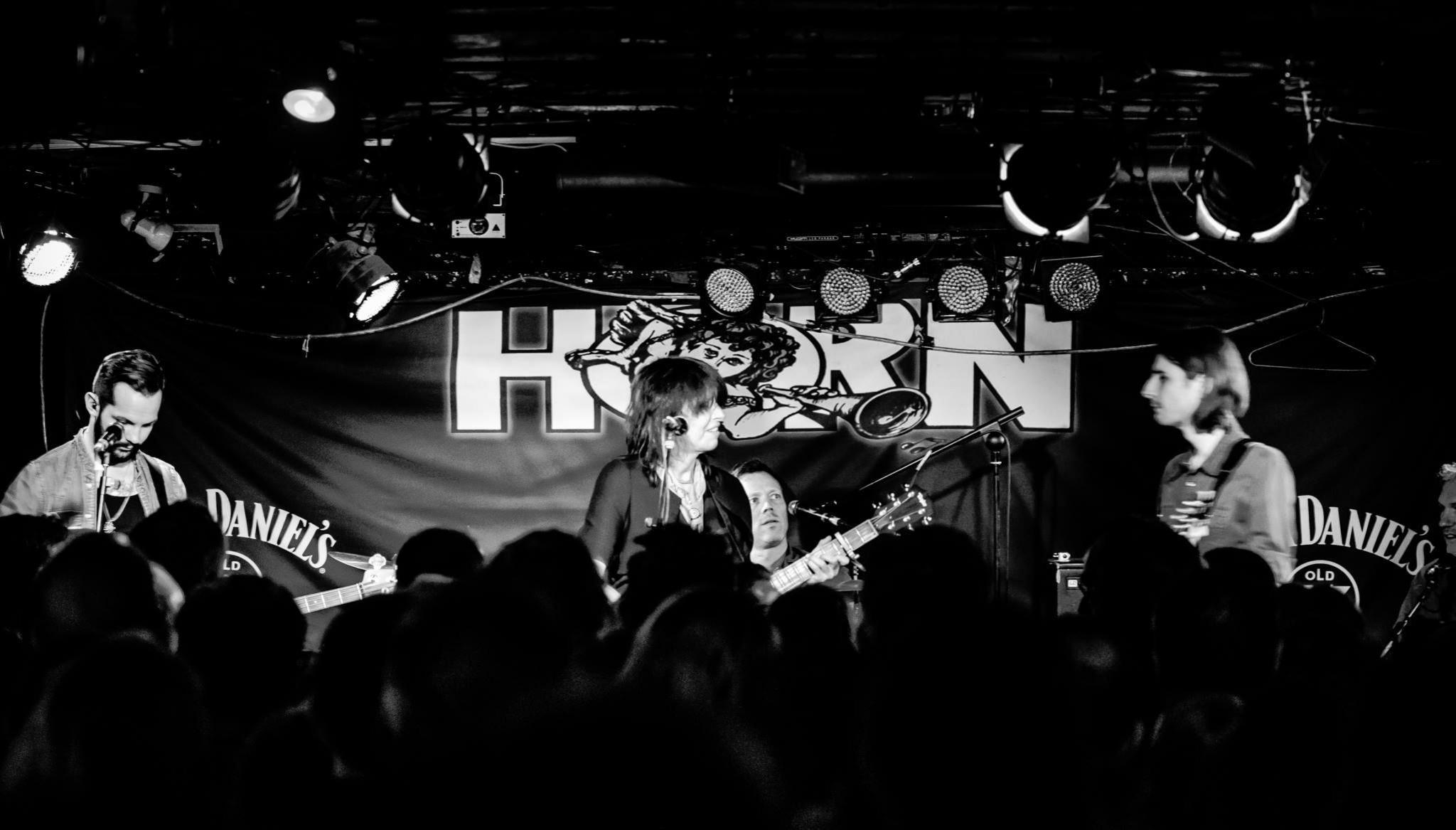 Chrissie Hynde performing at The Horn. The black and white image shows a female singer in the middle, holding a guitar, by a microphone, with a drummer behind her, and a guitarist to her left and right. People are in front of her but they are blurred. Lights are above the performers. 