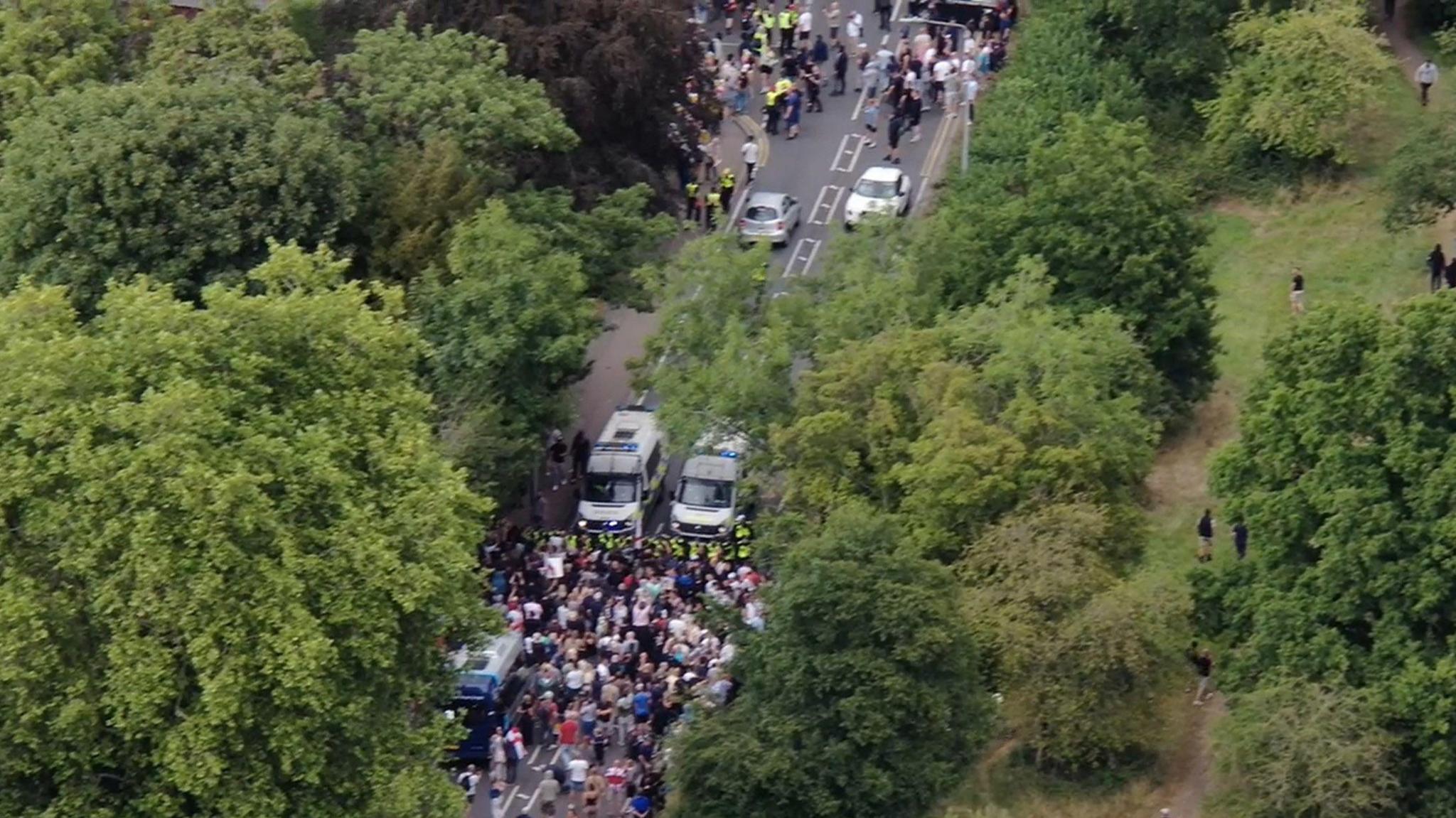 A drone image showing police using a van and a cordon to separate groups of protesters on a road, which is lined with large trees.