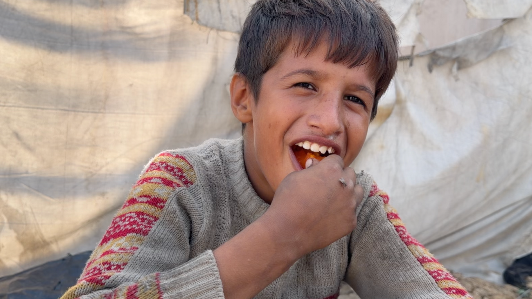A boy, with medium-length dark hair, bites into some food in his hand. He looks happy with his mouth open wide. Behind him is what looks like a white canvas tent and he is wearing a grey knitted jumper with yellow and red decoration on the tops of the sleeves.