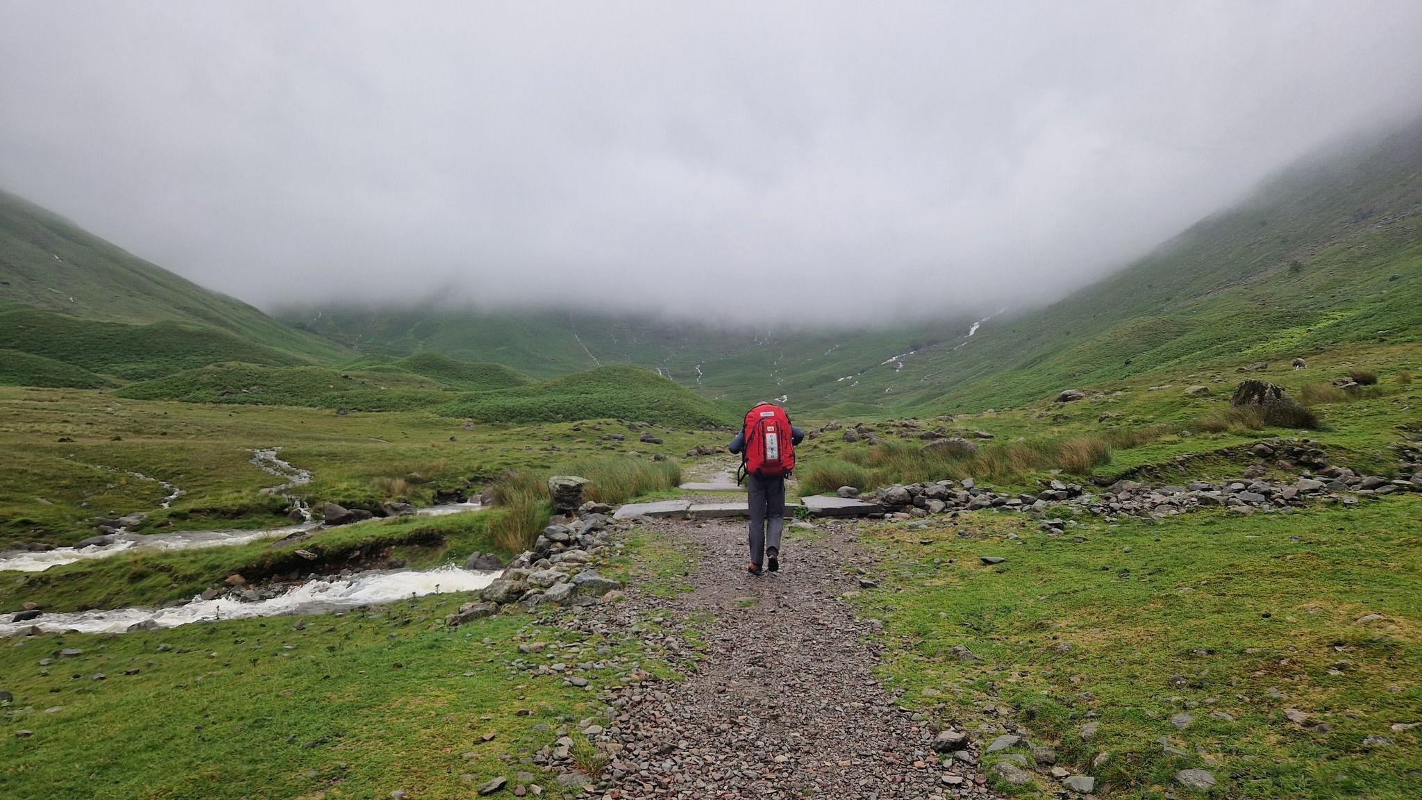 A mountain rescue volunteer, wearing a large red backpack, walks into the distance. There is a huge crag and fog in the distance, covering the Lake District fells.