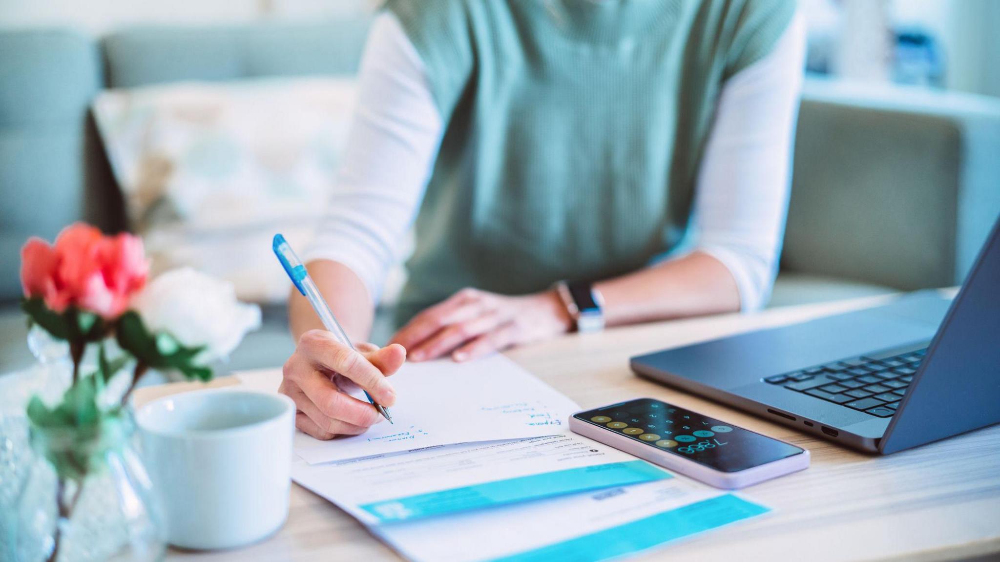 A stock image of a woman holding a pen with writing on some forms. In front of her is a laptop and an iphone which is open on the calculator app. To the left are some flowers in a vase and a mug.