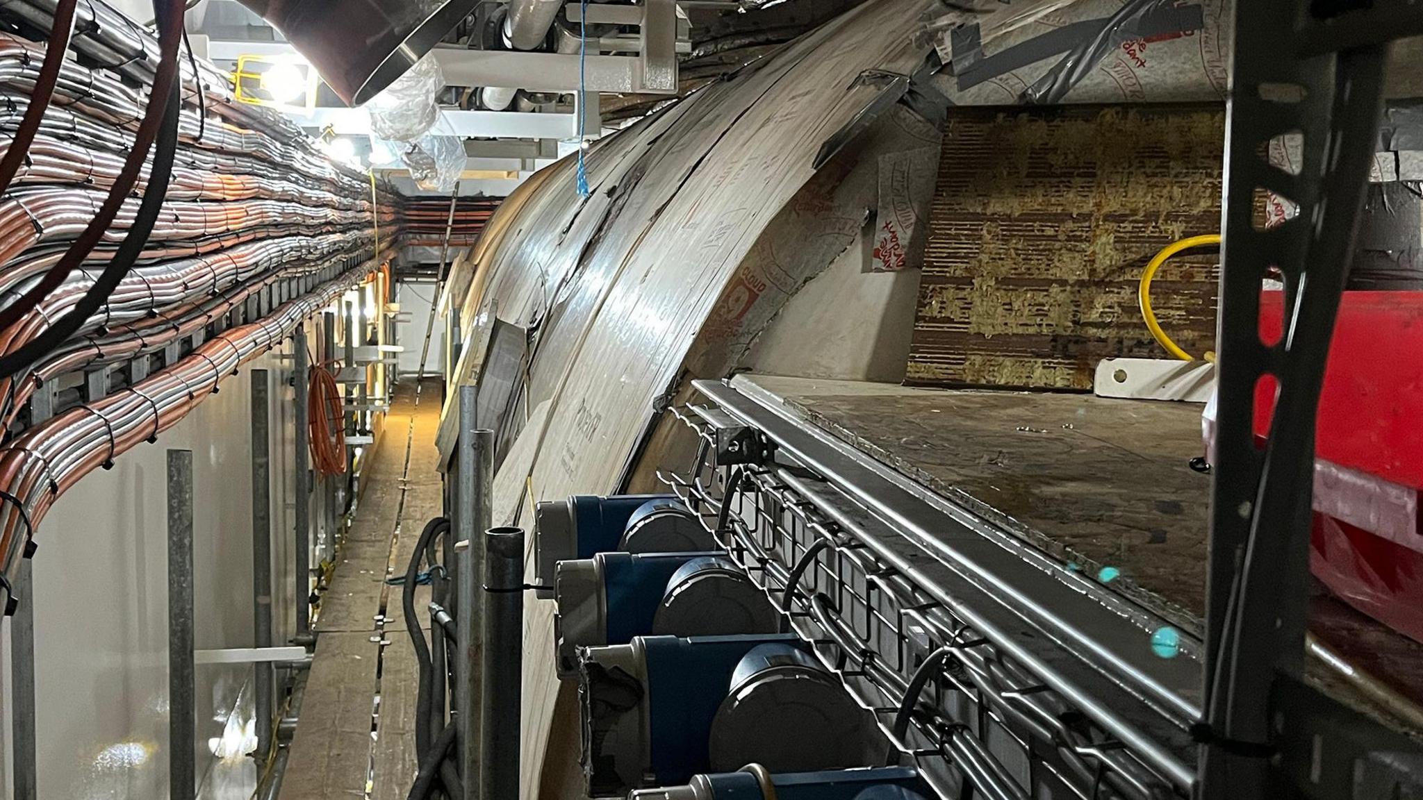 A large cylindrical  tank inside a ship with various pipes and cables running along the left side of the picture