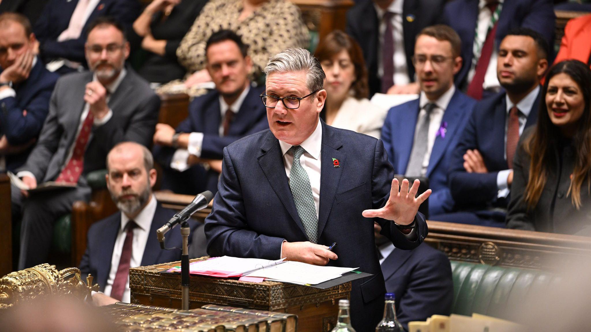 Sir Keir Starmer, wearing dark rimmed glassed, a navy suit, white shirt, light blue tie and a poppy, addressing the House of Commons at the despatch box with Labour MPs behind him.