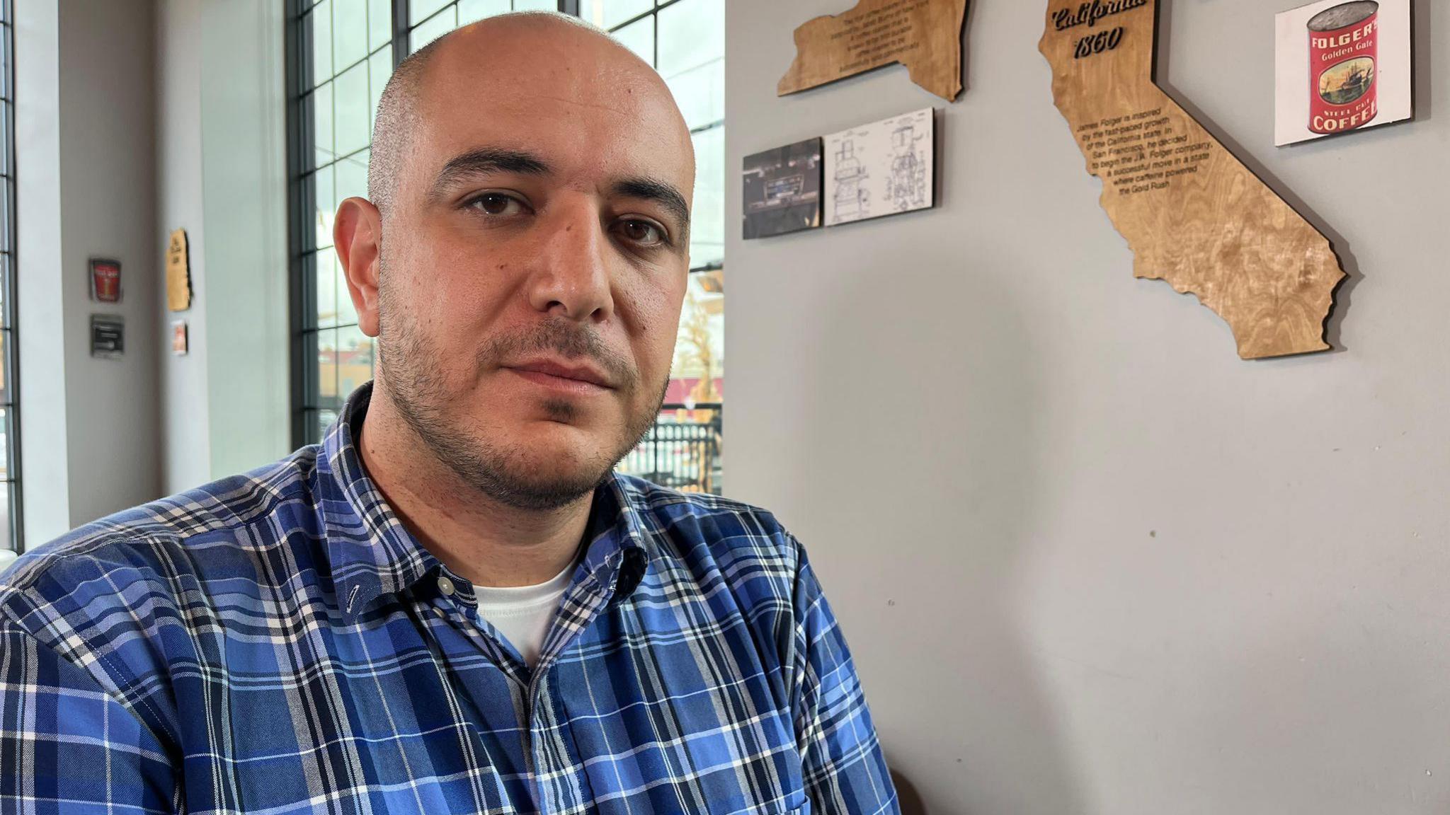 Abbas Alawieh sits in a blue plaid shirt in an office-like setting. There is a large window behind him.