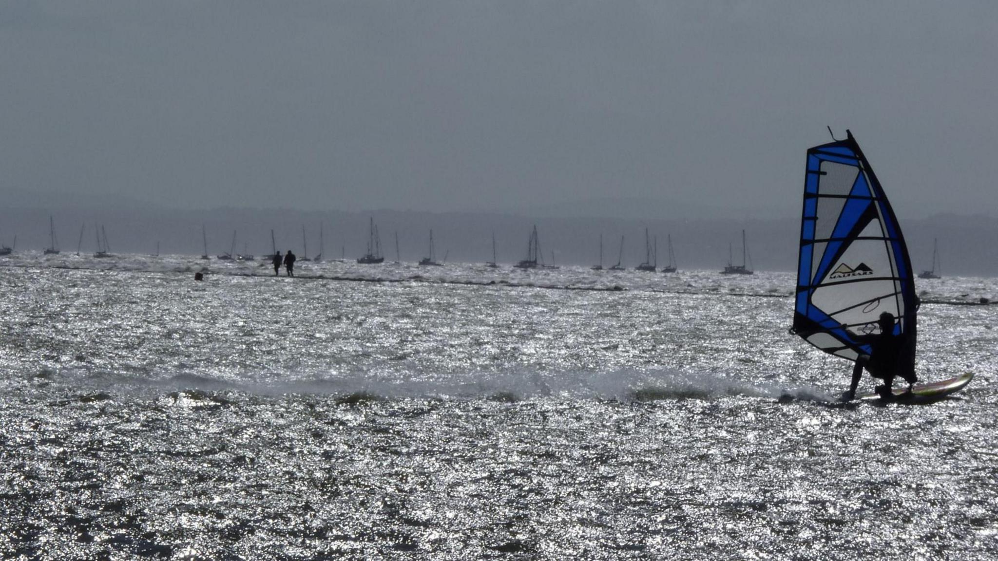 A windsurfer glides on the gentle waves of West Kirby, with various boats and yachts in the sunlit background