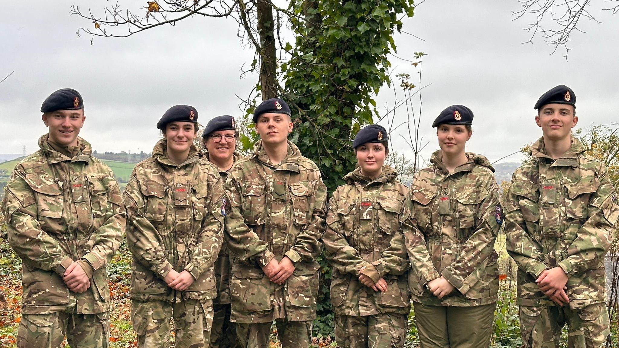 One woman, three girls and three boys wearing camouflage uniform with black cadets hats stand outside. 