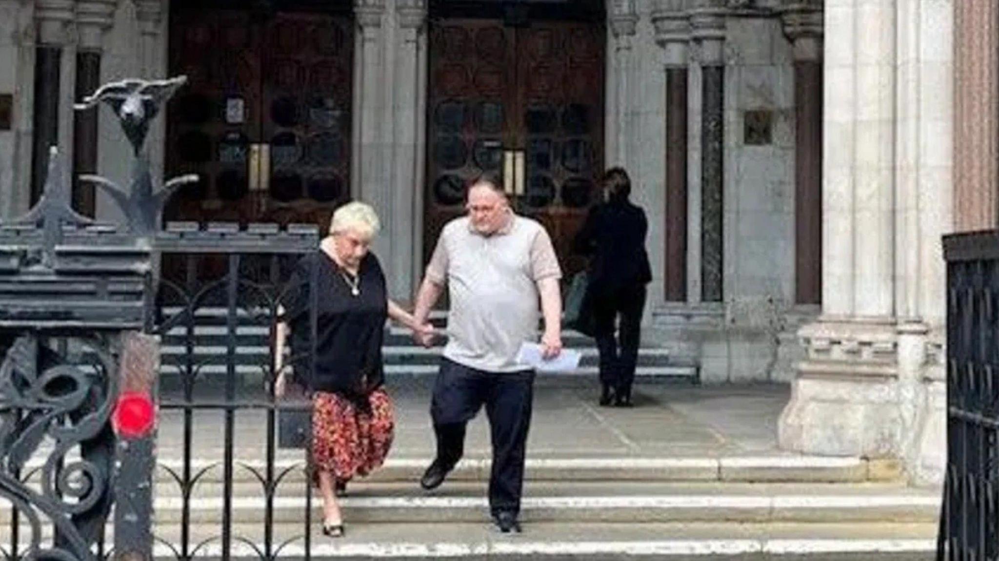 Ann Ming, wearing a black top and red skirt, walks down the steps of the Courts of Justice with Kevin Hogg, who is wearing a grey polo shirt and black trousers.