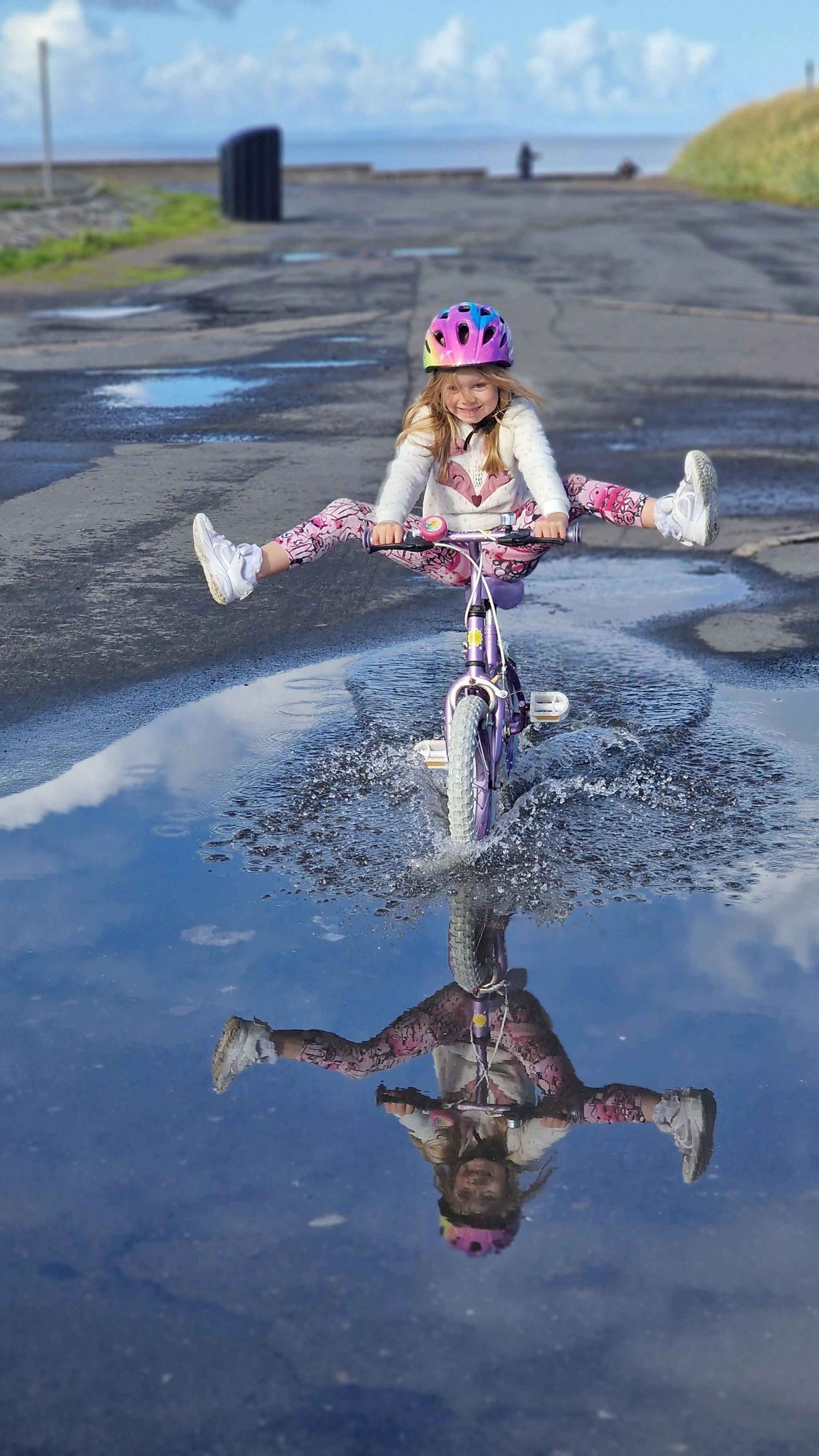 A young girl in a multi-coloured outfit and cycle helmet cycles through a puddle near the waterfront in Girvan, her legs lifted up and freewheeling through a puddle