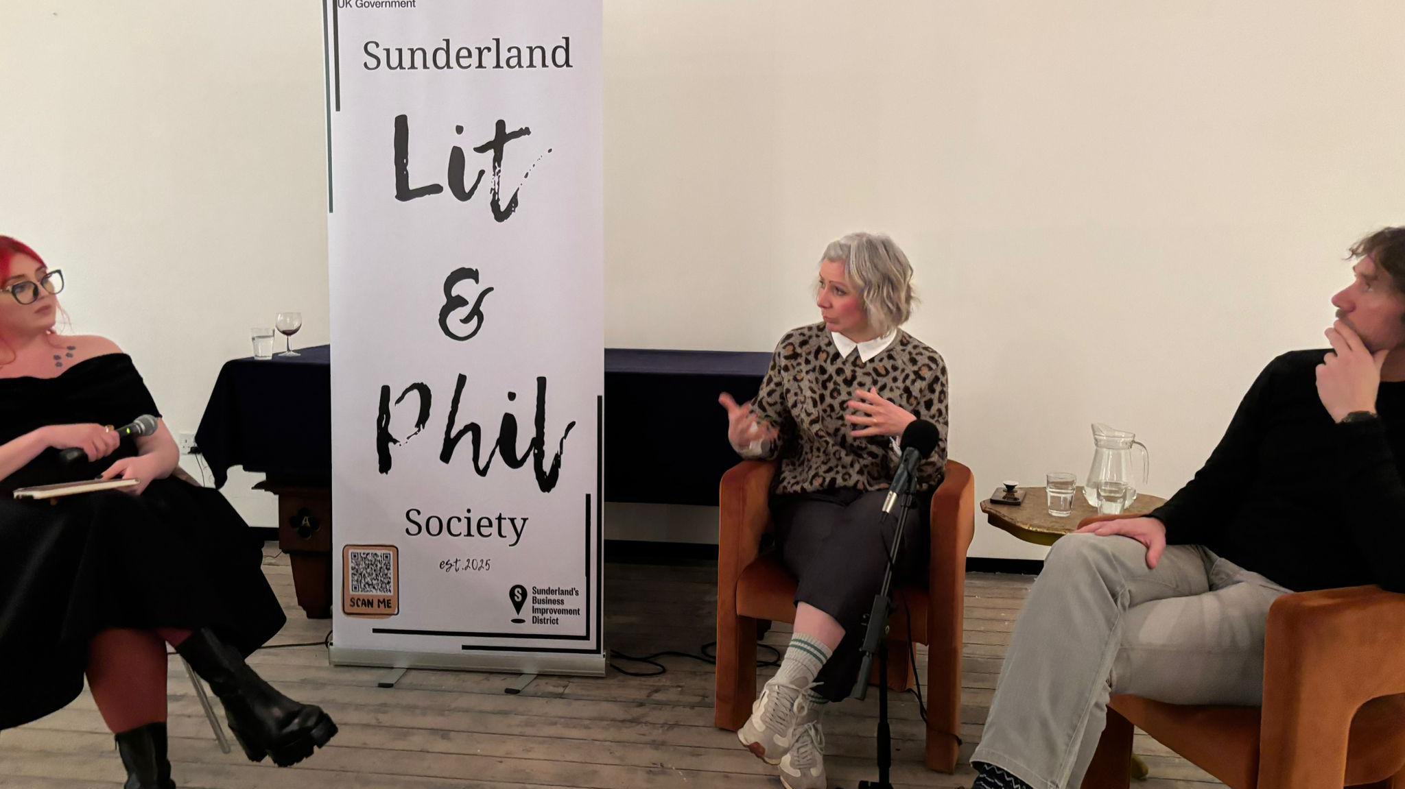 Three people sitting beside a Sunderland Lit and Phil Society banner. A woman with light hair is talking to Ms Langley to the left who has red hair. A man can be seen listening to the right.