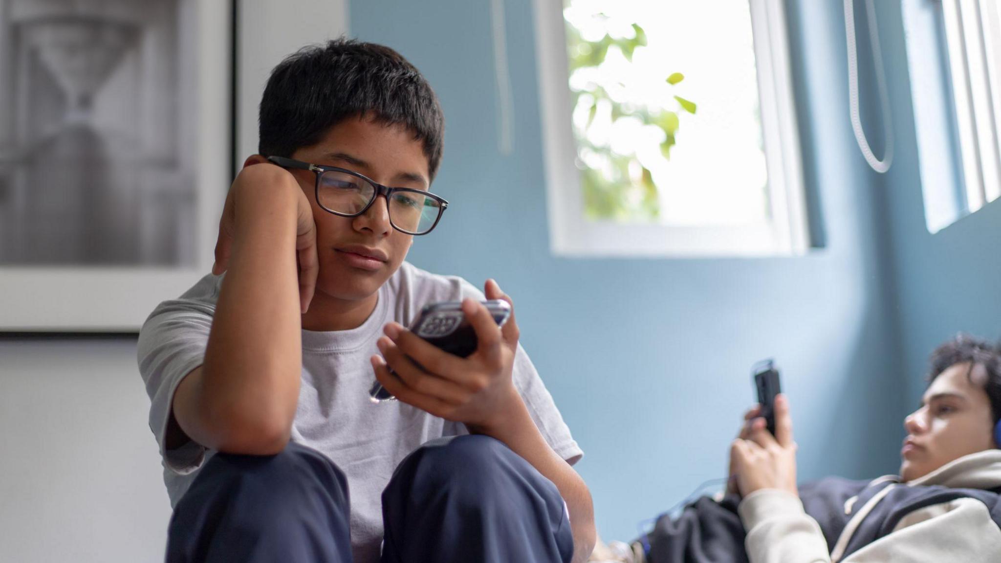 a kid wearing glasses looking at his phone with another kid out of focus in the background lying down also looking at his phone