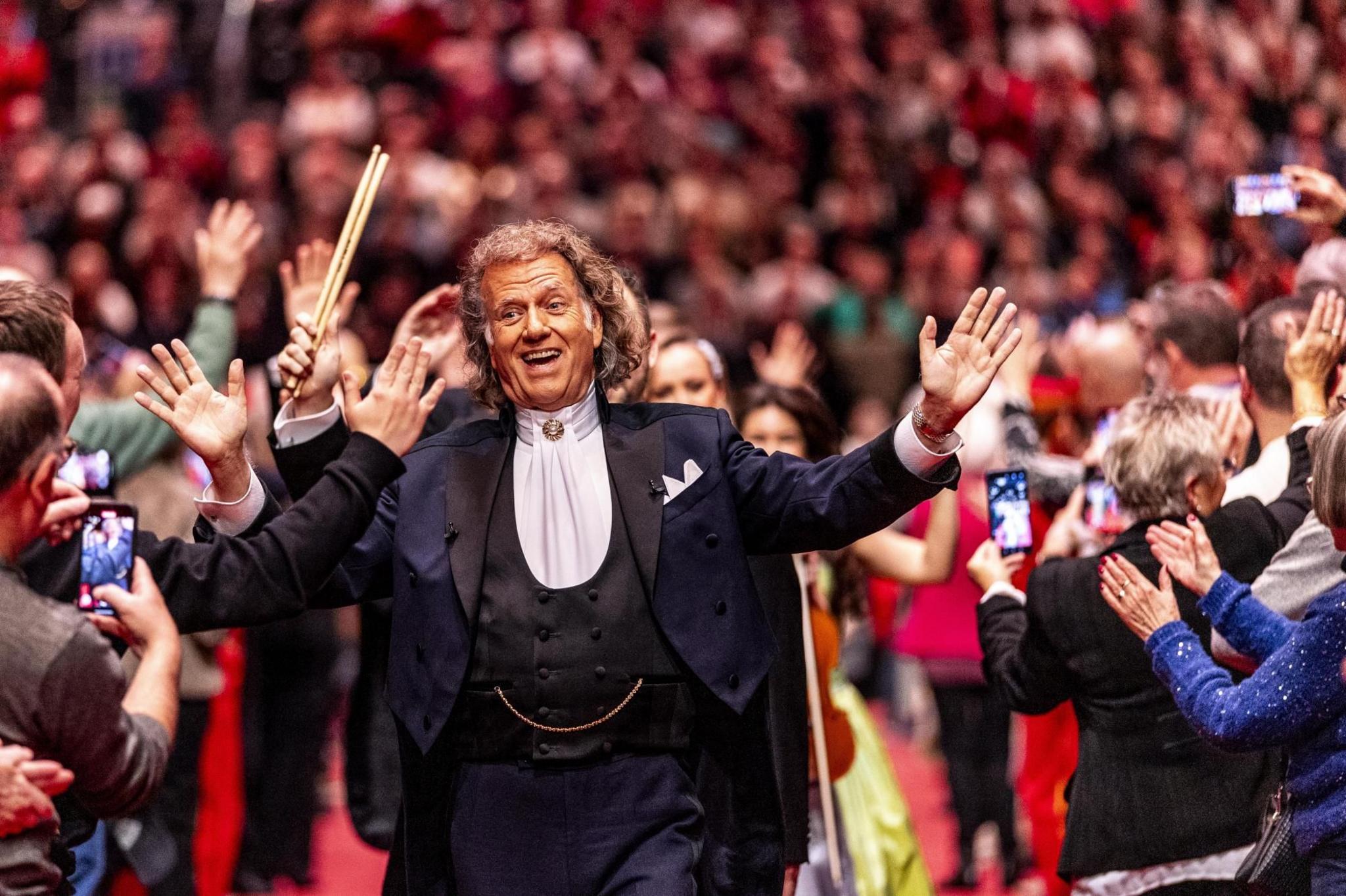 Violinist Andre Rieu, dressed in a black suit with white shirt, walks, smiling, arms wide apart, through a crowd of people, some taking photographs with mobile phones, in Maastricht, in the Netherlands.
