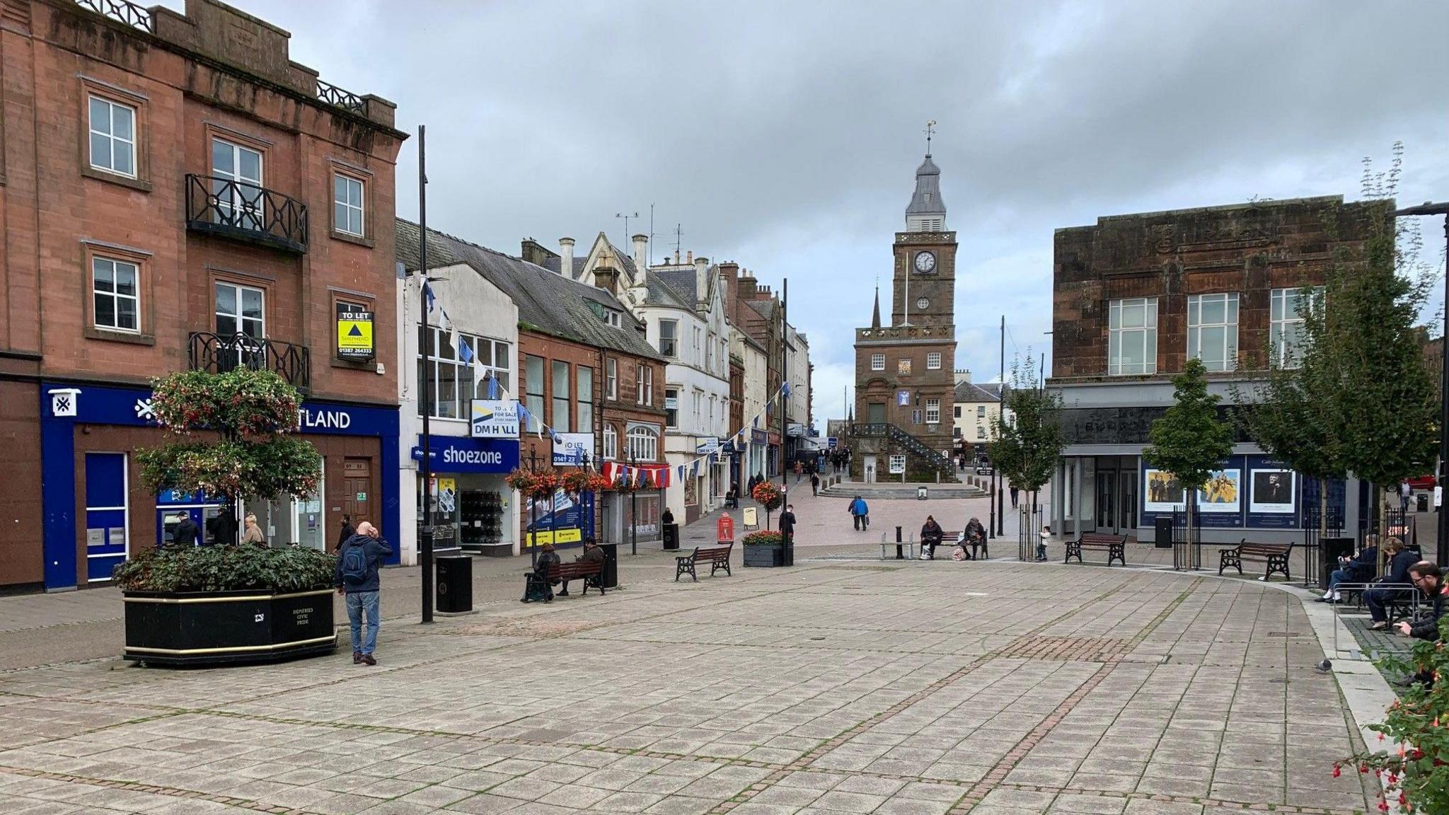 A town centre with a series of shops, benches and a few people milling about with a large sandstone steeple building in the distance