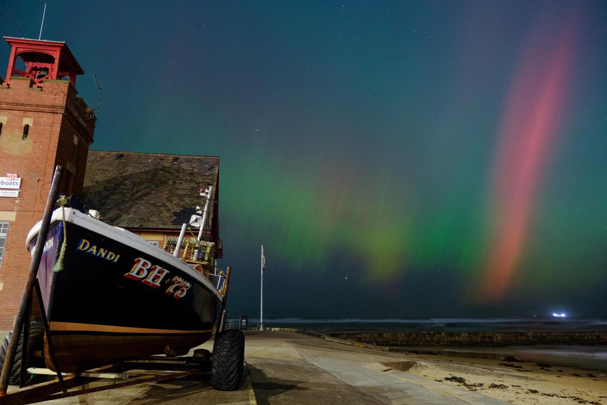 A small boat on a trailer is parked on the left-hand side of the image, Behind it is Cullercoats lifeboat station, a brick building. The sky has red and green streaks of light.