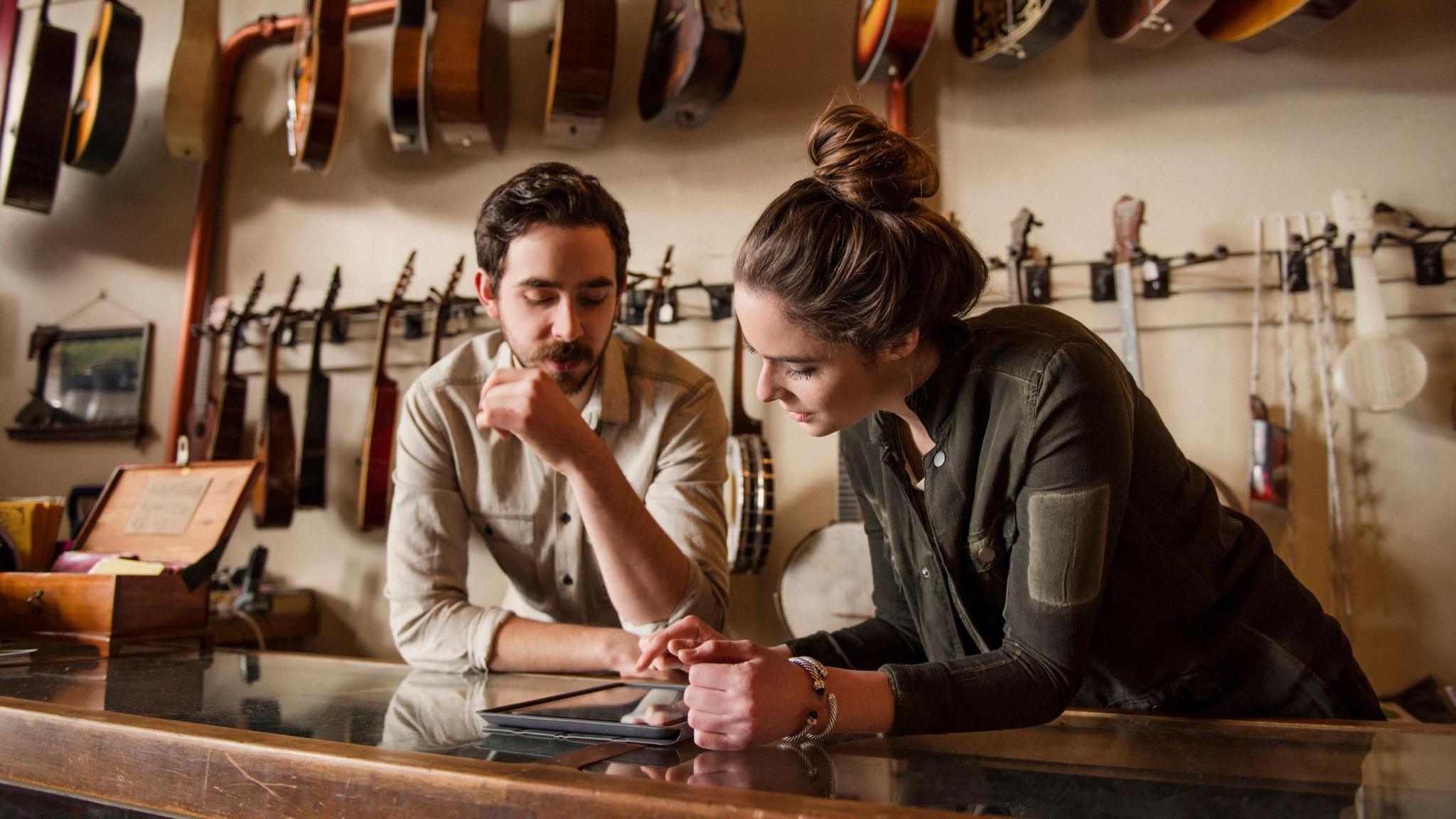 A young couple, a man and a woman, standing behind a wood and glass counter in a music shop. They are looking down at tablet device presumably displaying information about their business. Two racks of guitars are hanging behind them.