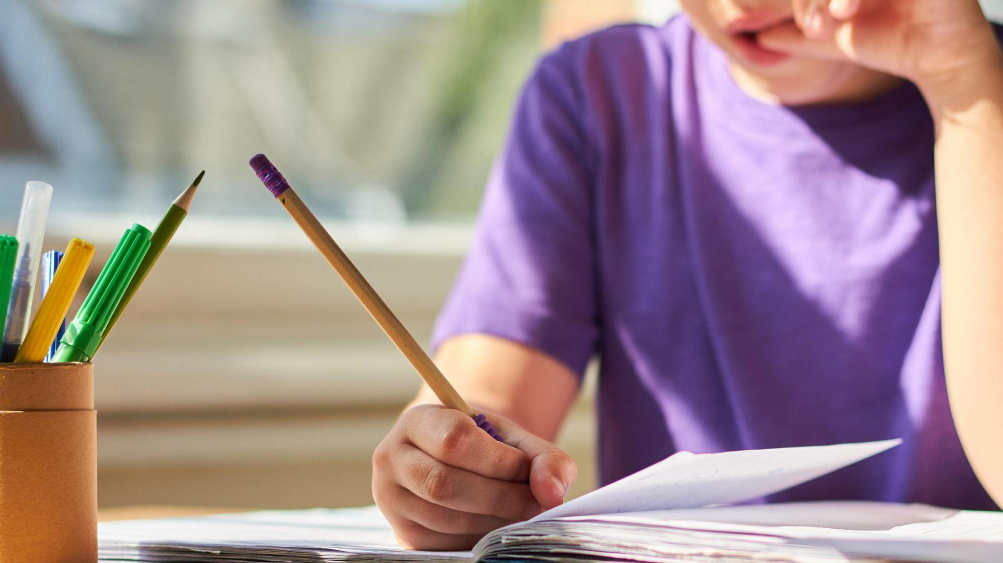 A stock image of a child in a purple T-shirt writing with a pencil