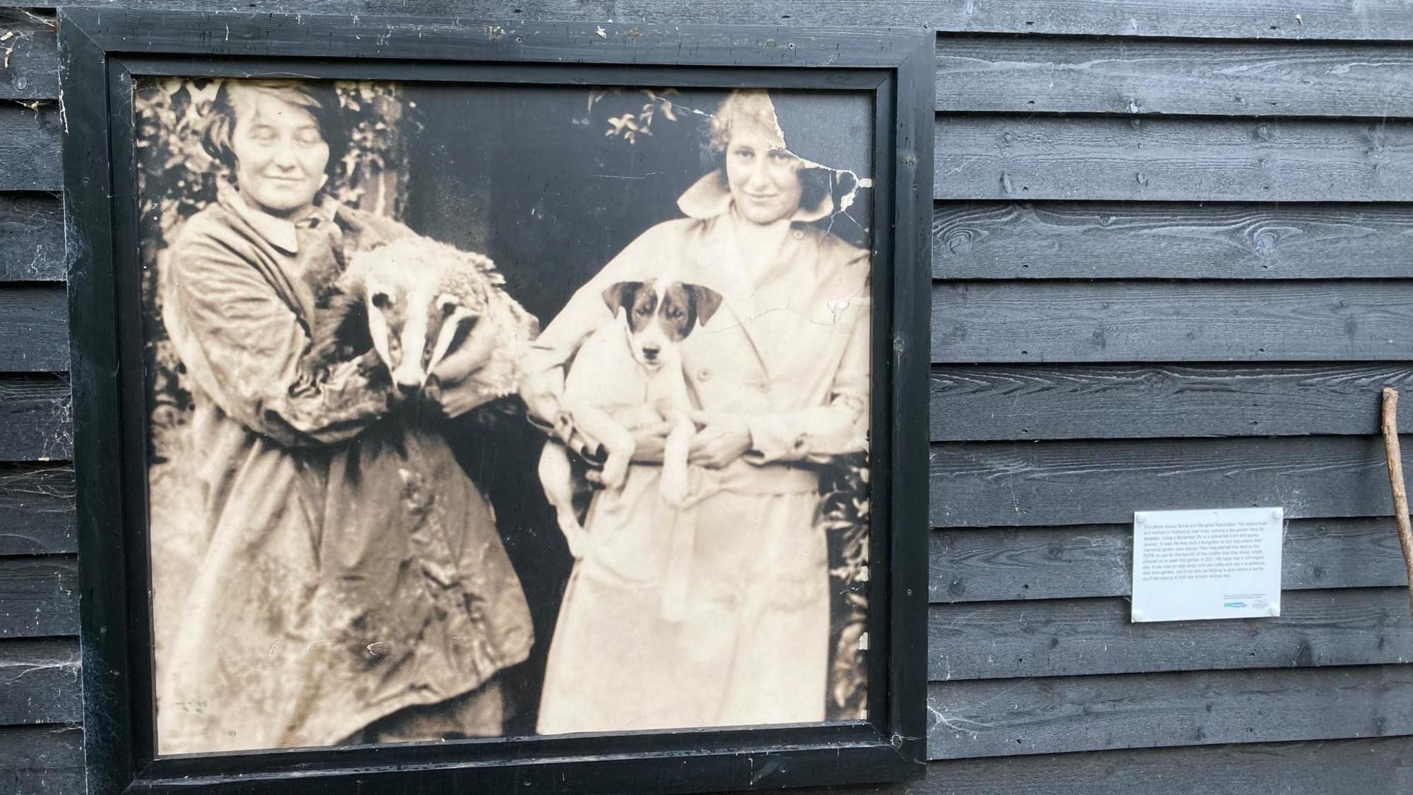 A black and white image of two women, one holding a dog, the other holding a badger, within a frame on the exterior of a black, wooden-clad building. The is a small information sign next to the picture.