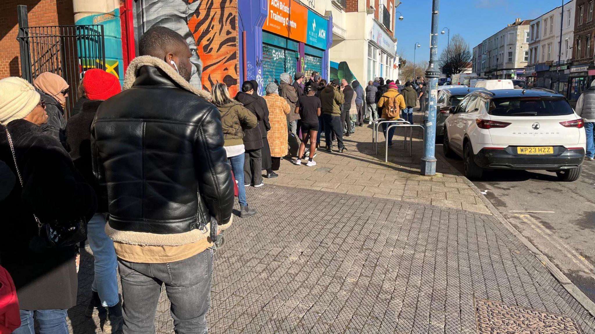 A long line of people is seen queuing around a building in St Paul's in Bristol as they wait for the chance to register at a dental surgery. They are wearing warm clothes as it is a cold day, although it is clear and sunny