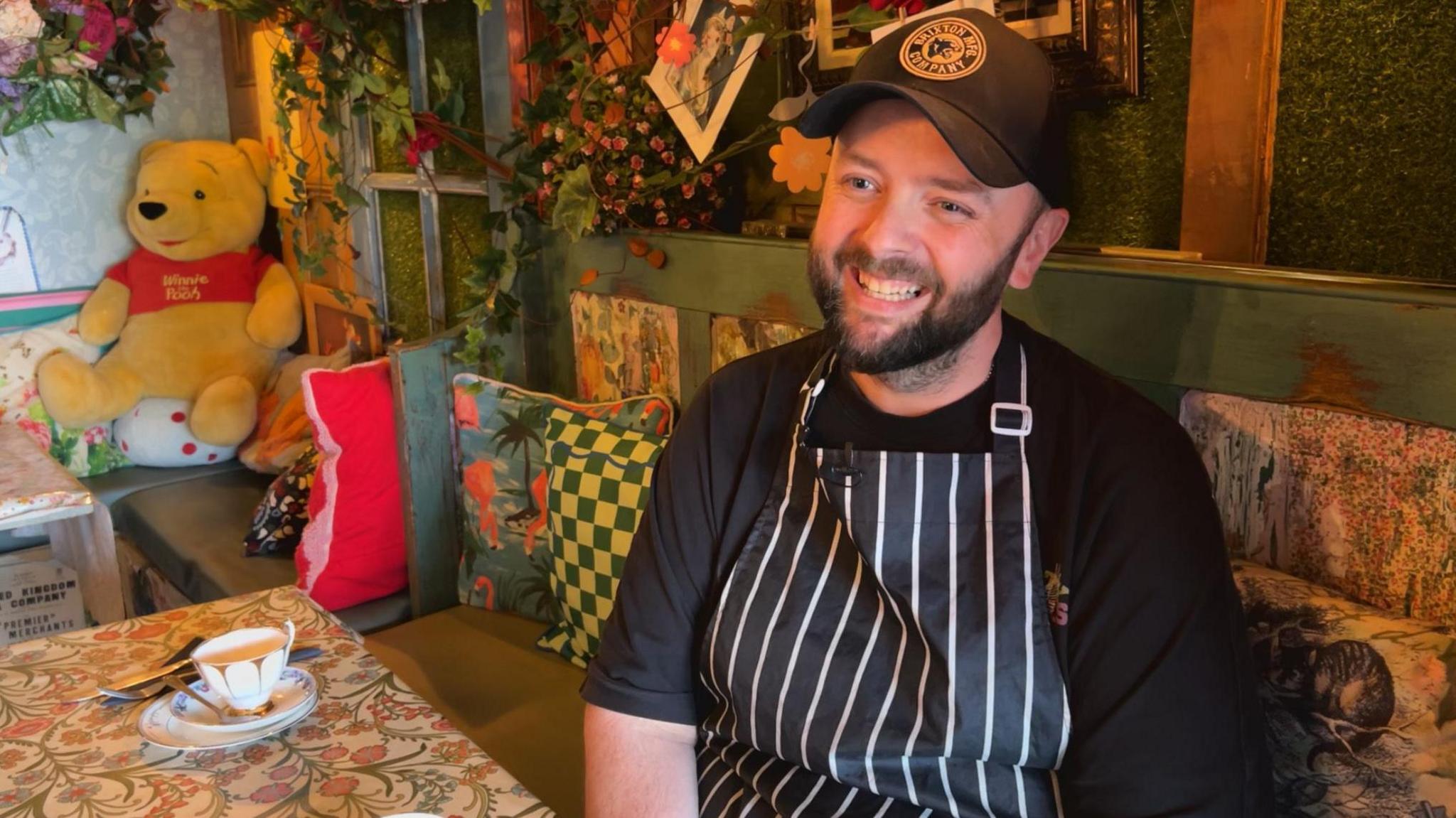 A man wearing a striped apron and cap seated in a café decorated with plants, patterned cushions and a Winnie the Pooh toy.