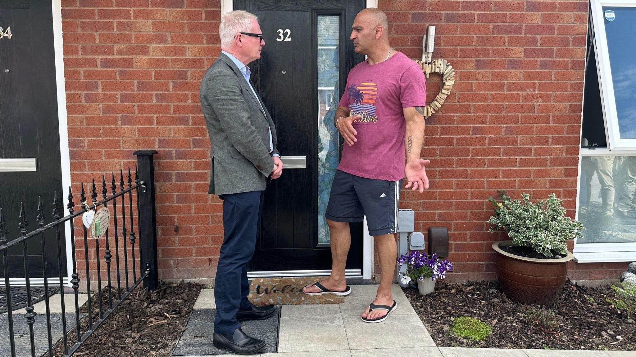 Man in a green suit jacket standing on a doorstep talking to a man in a t-shirt, flipflops, and shorts