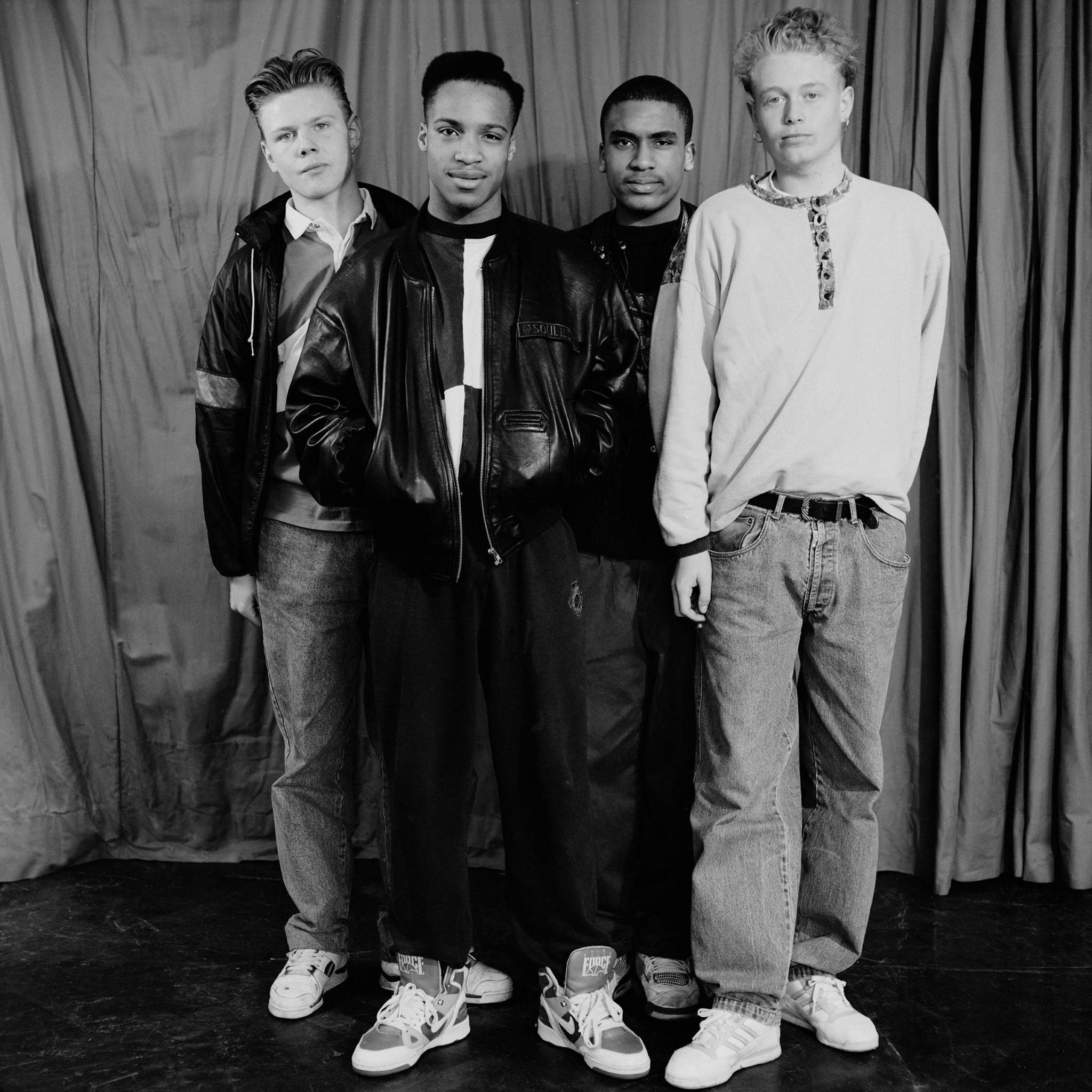 Boys from Tulse Hill School, 1990. Four young men stand in front of a curtain. They wear jeans or tracksuit bottoms, trainers and casual tops. Three wear leather jackets. Black and white photo.