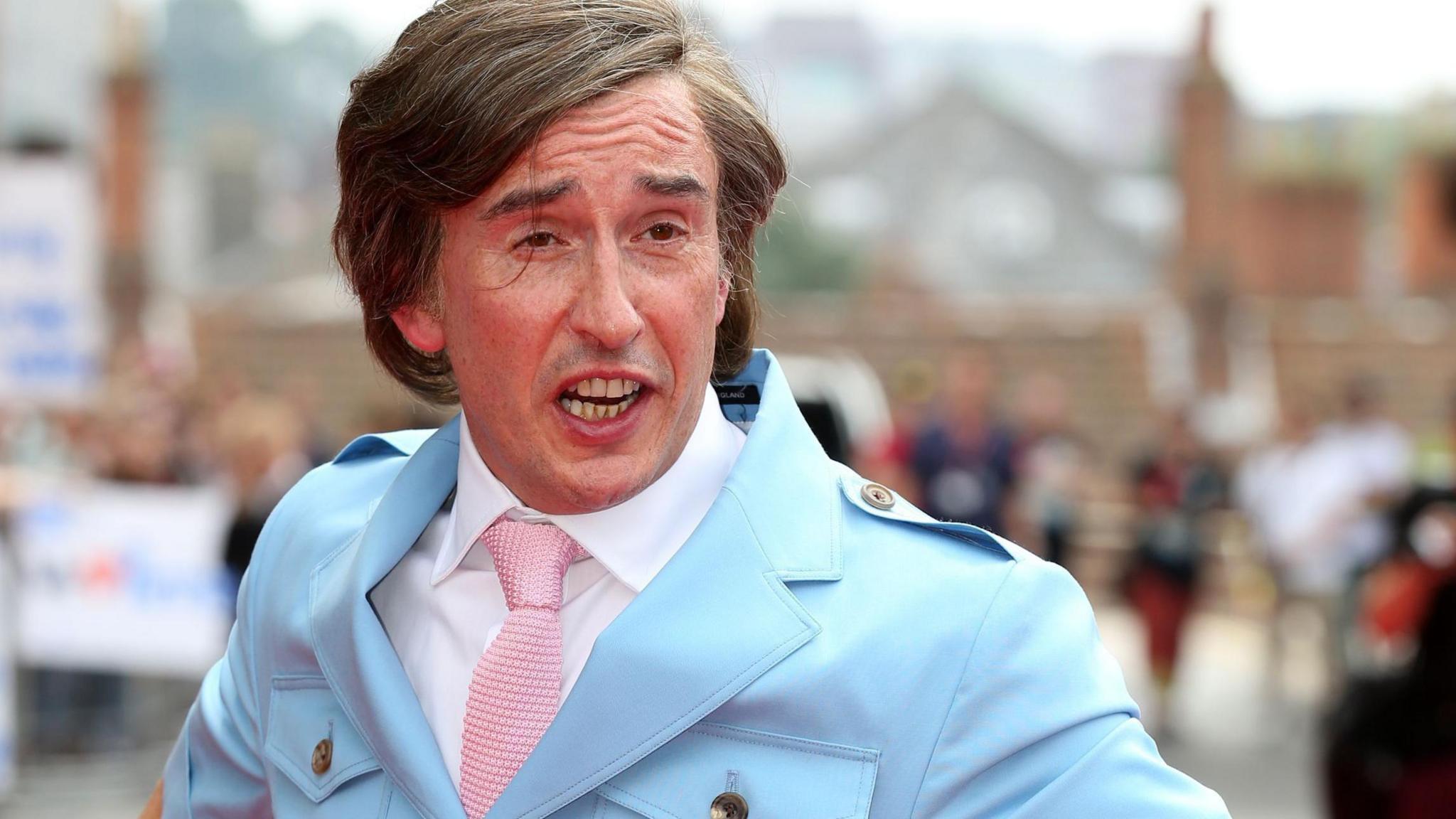 Steve Coogan as Alan Partridge, with a sweeping fringe and dressed in a light blue safari suit with a crisp white shirt and baby pink tie. He is outside with blurred crowds behind him.