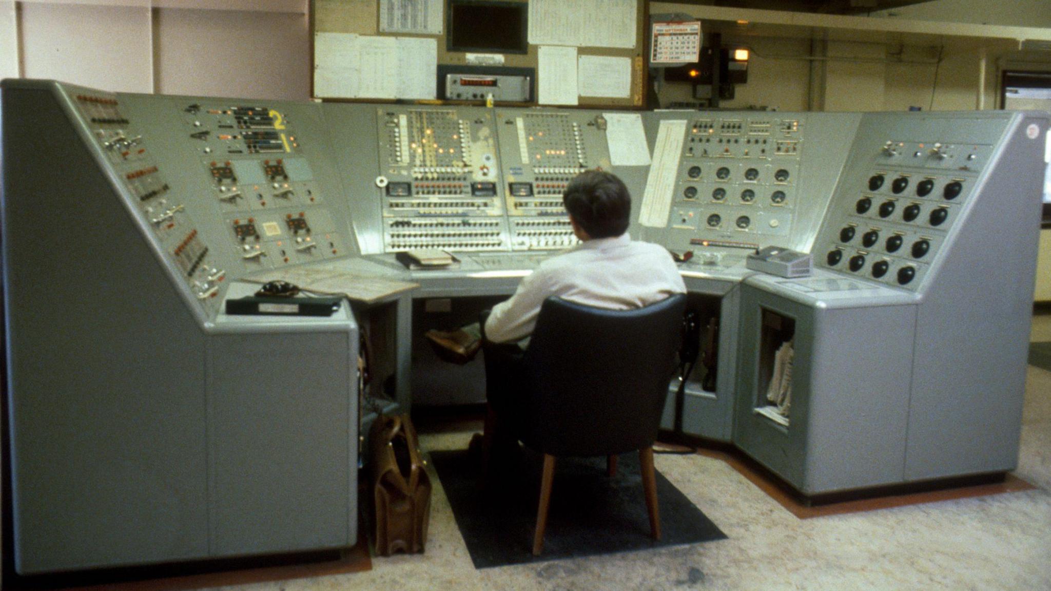 Person sitting in a black chair looking at a semi-circular grey console with dials, and switches in several groups on the six panels in front of him. There is equipment set into the wall beyond the console.