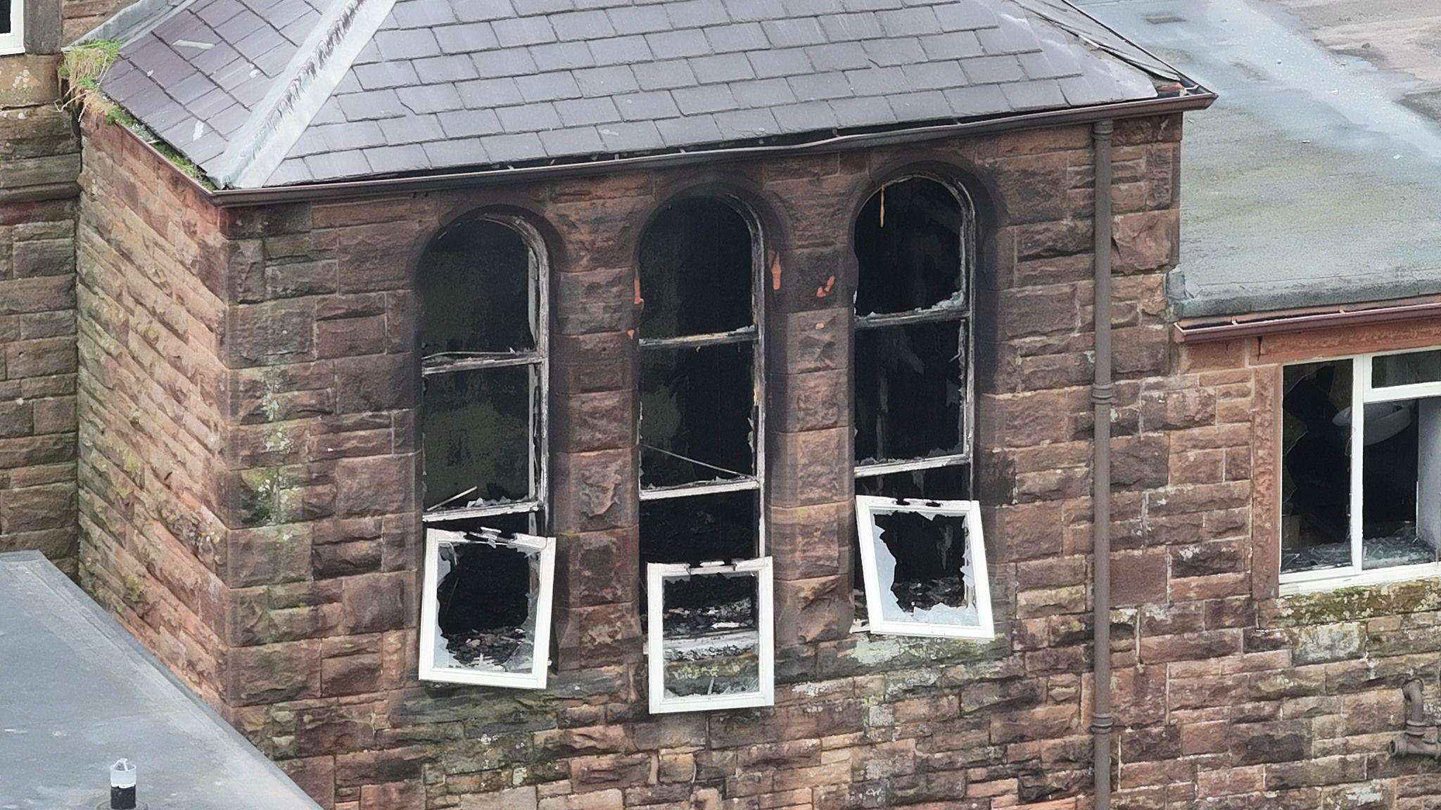 Burned-out windows on an old sandstone building with a slate roof. The window panes have all fallen out apart from three which are hanging on their hinges.