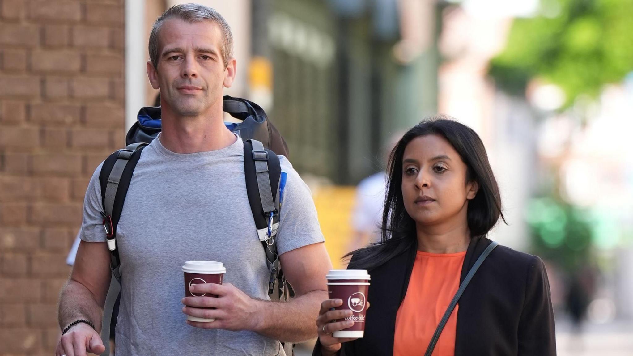 Sean and Shiza Harper outside Chelmsford Crown Court. They are holding coffee cups. Sean Harper looks to camera. He has greying hair and is wearing a grey t-shirt and has a large rucksack on his back. Shiza Harper has shoulder-length dark brown hair. She is wearing a dark jacket with an orange top under it.
