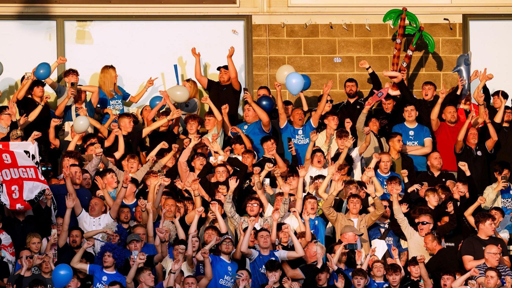 Football fans with balloons and inflatables sing in the sunshine while standing on the terrace.