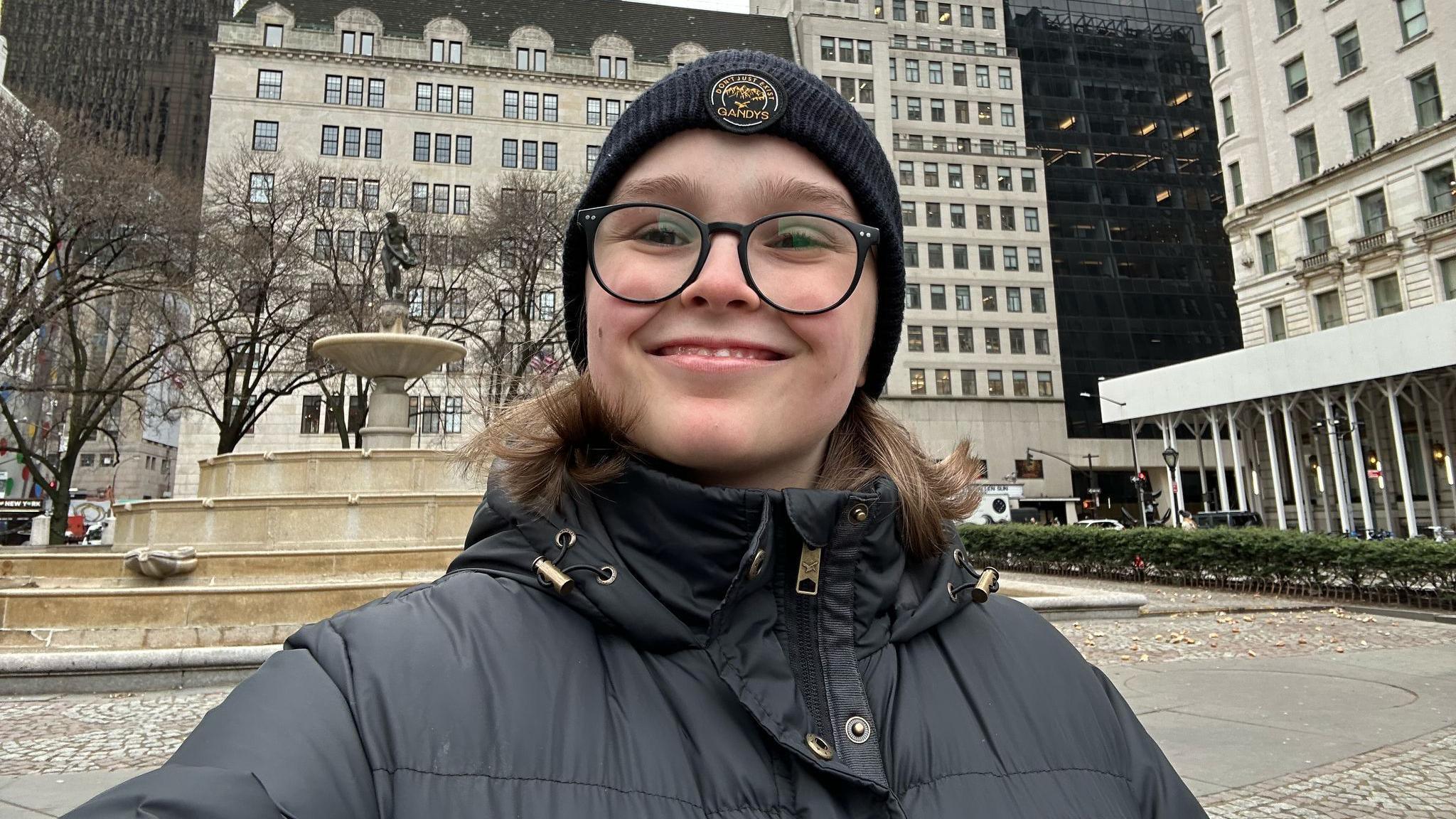 Bethan stands in front of a fountain. She smiles broadly to camera and is wearing a black puffy coat, a black beanie and black round glasses.