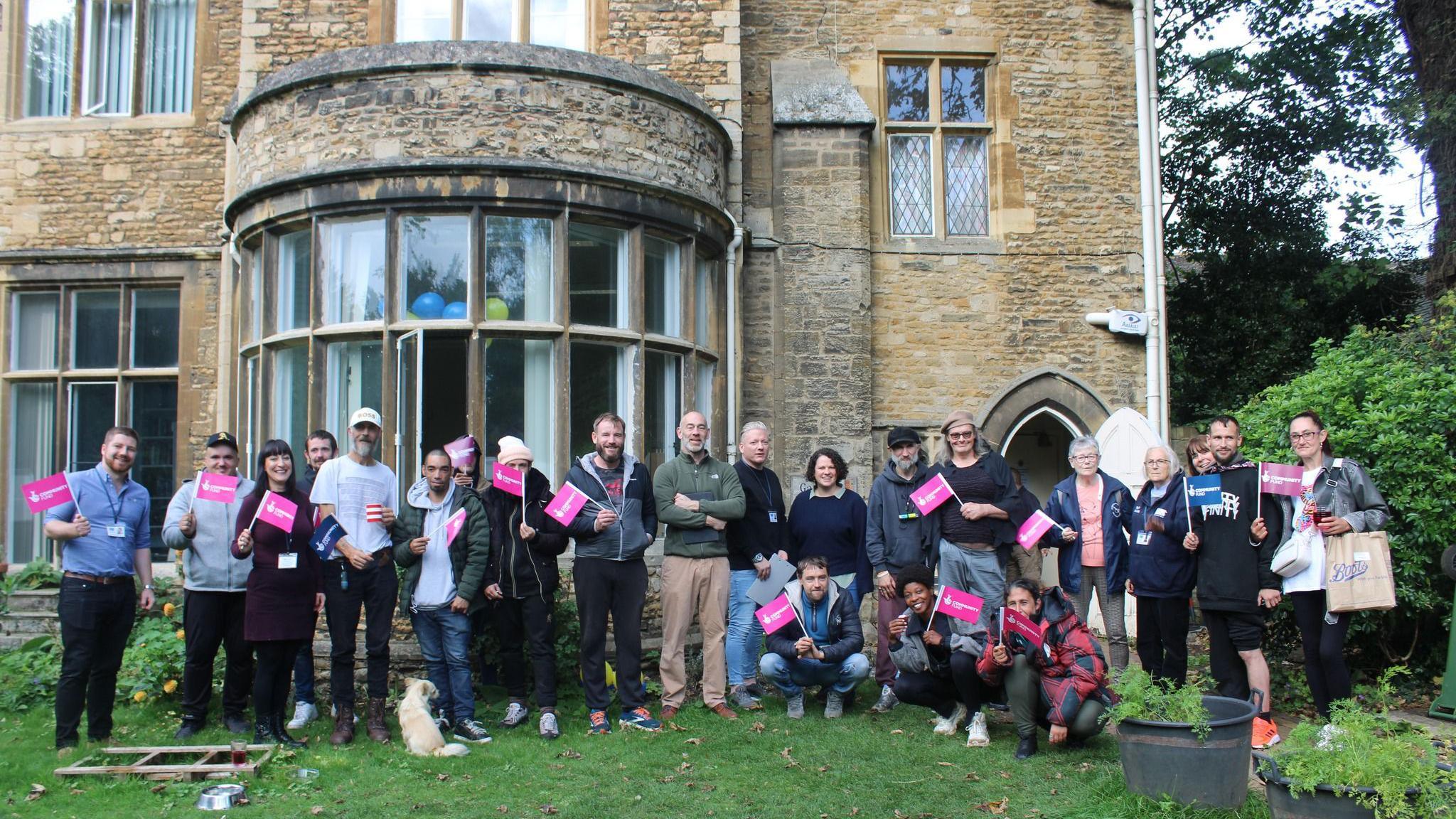 Volunteers and users of Light Project Peterborough's Garden House, standing in line on the grass in front of the grand stone building. They are holding pink small flags that say "National Lottery". 