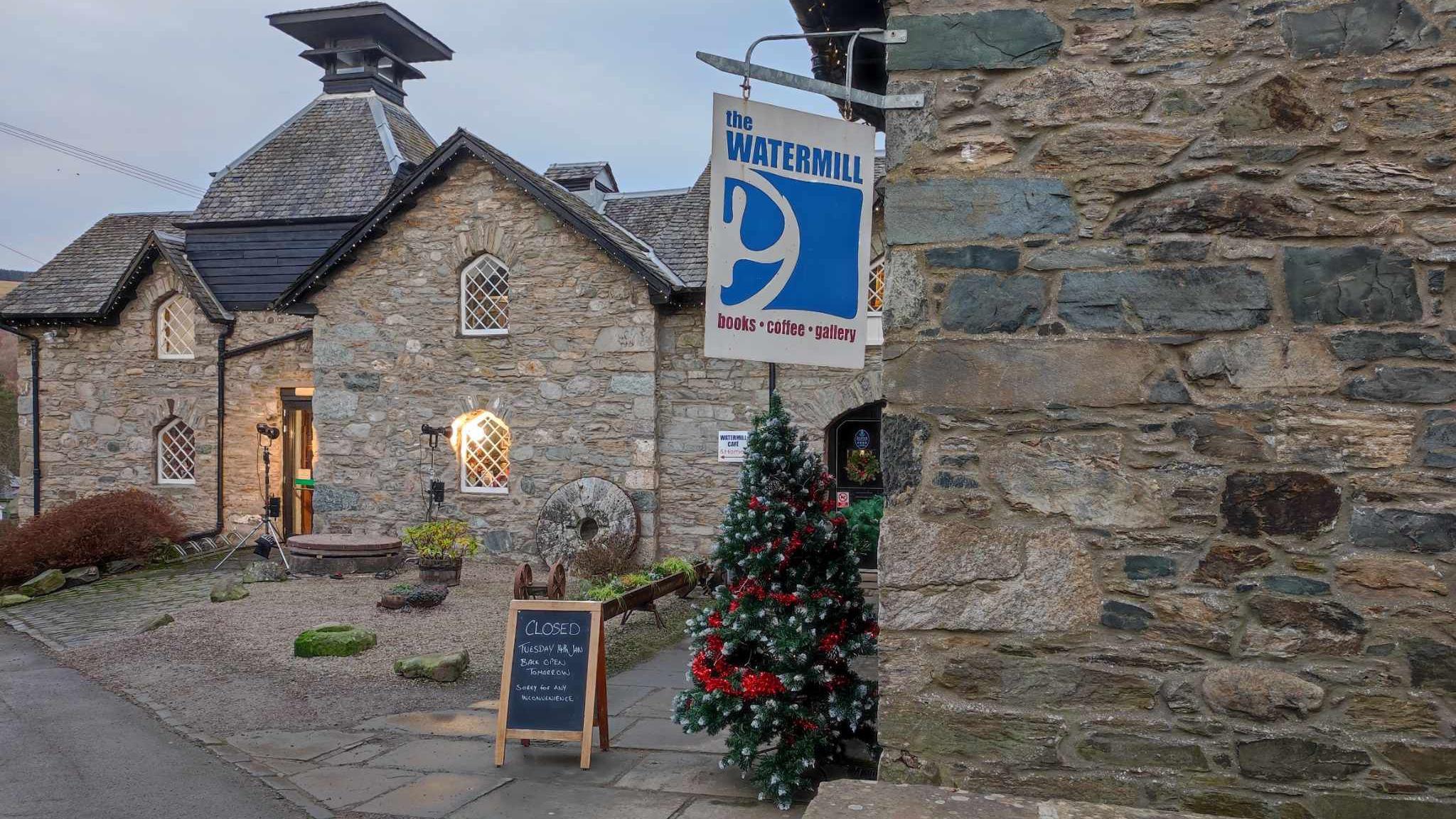 A stone building is decorated with a Christmas tree and a sign hangs from the wall that says The Watermill.