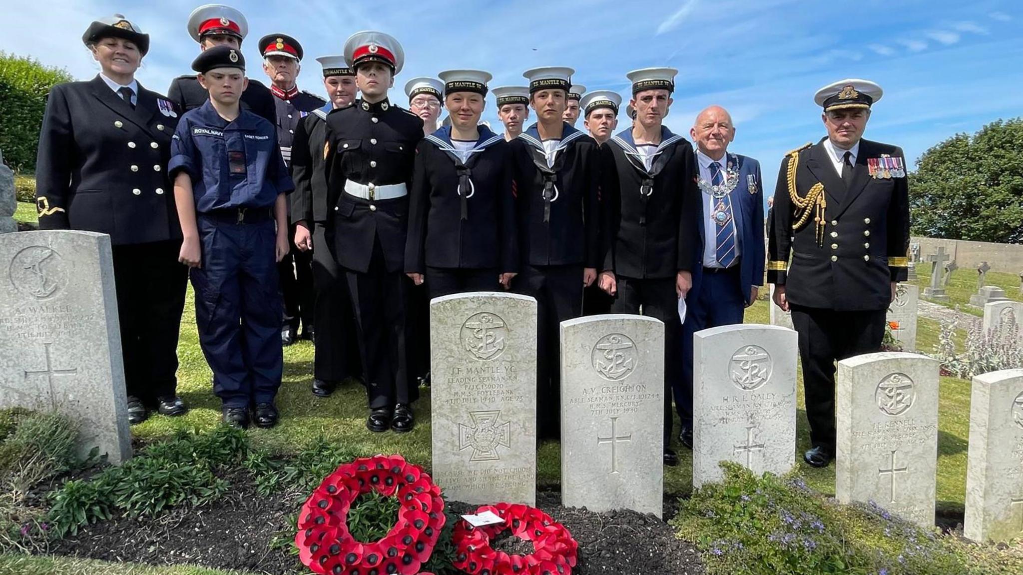 Dignitaries stand at the grave of Leading Seaman Jack Mantle on the 85th anniversary of his death during a German dive bomber attack on Portland Harbour on 4 July 1940. They included HM Lord-Lieutenant of Dorset Michael Dooley, the Mayor of Portland Town Council Charlie Flack, Portland Sea Cadets and Yeovil Sea Cadets from TS Mantle VC, named in honour of Jack Mantle. There are poppy wreaths laid at the grave. It is a clear day.