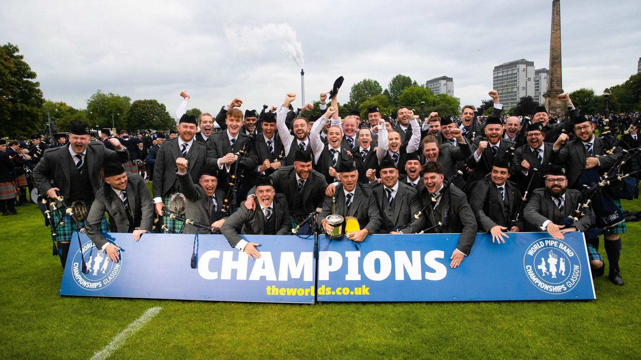 Members of a pipe band celebrate being crowned world champions. About 20 members dressed in kilts and jackets look happy posing behind a blue champions banner.