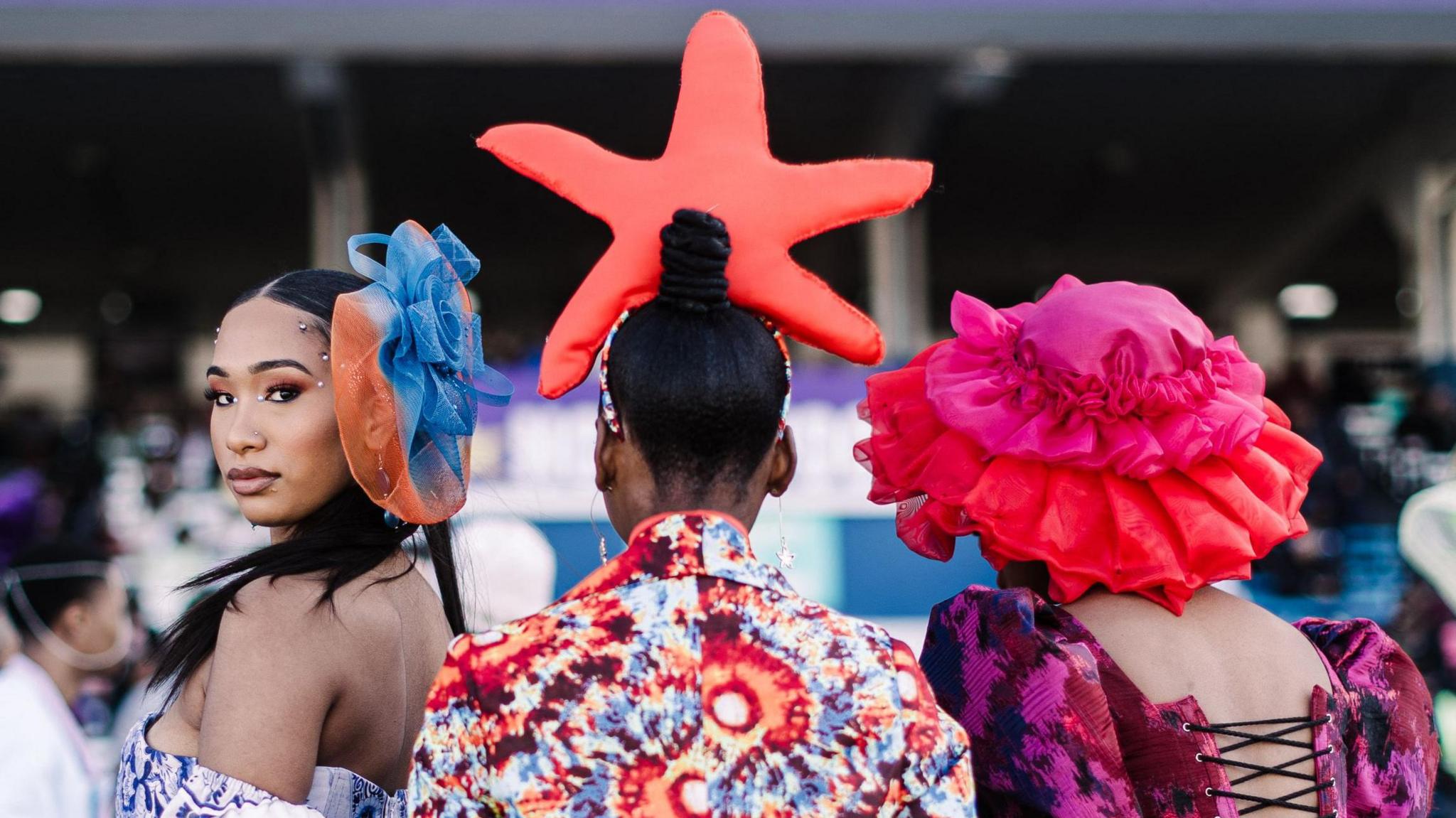 Women wear dresses and bold hats in bright colours. Their backs are to the camera, but one of them has turned to look over her shoulder towards the camera.