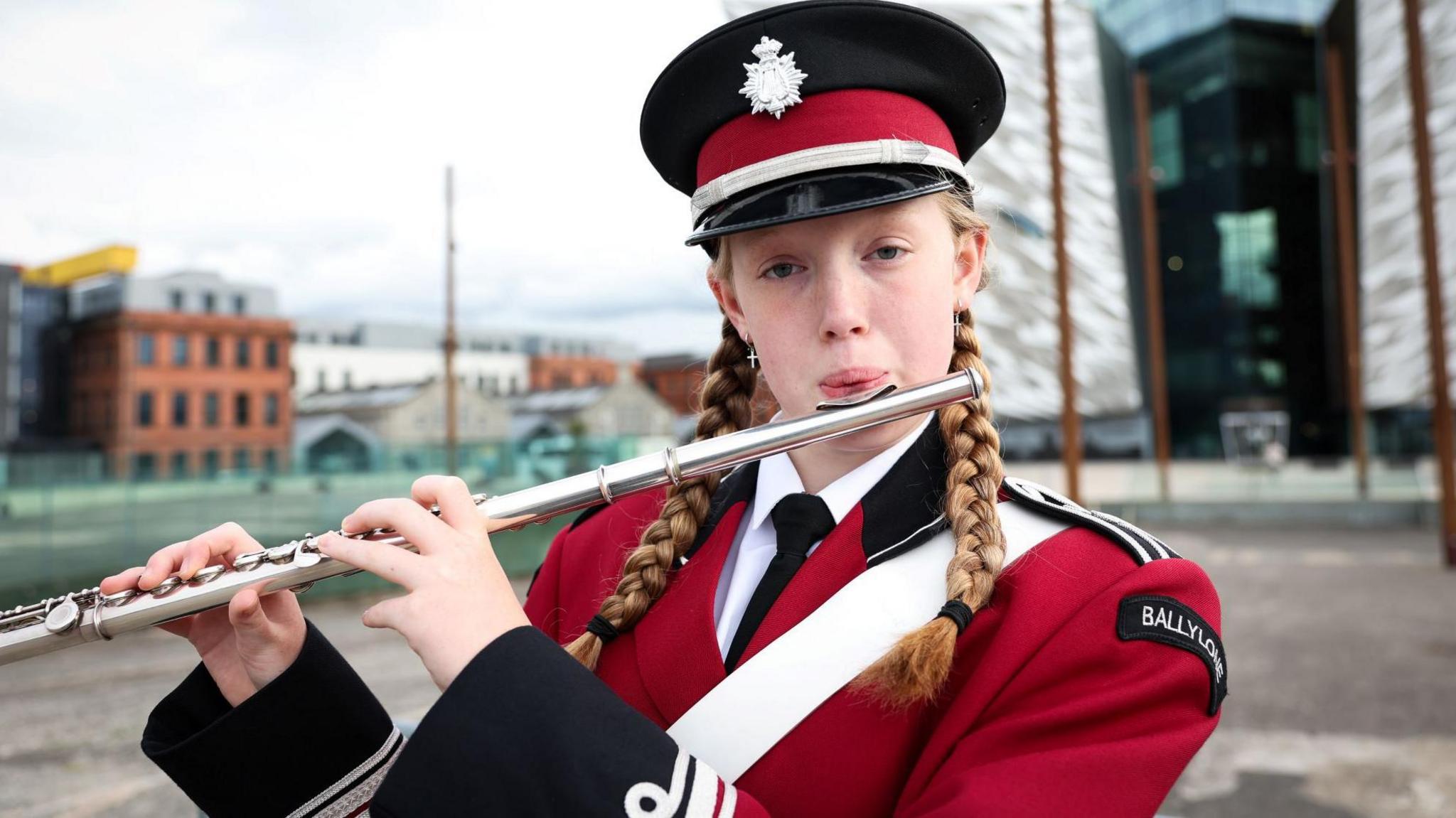 A girl is playing a silver flute. Her auburn hair is plaited. She's wearing a red and black band uniform. There's a silver medal attached to her cap. The Titanic museum is in the background. 
