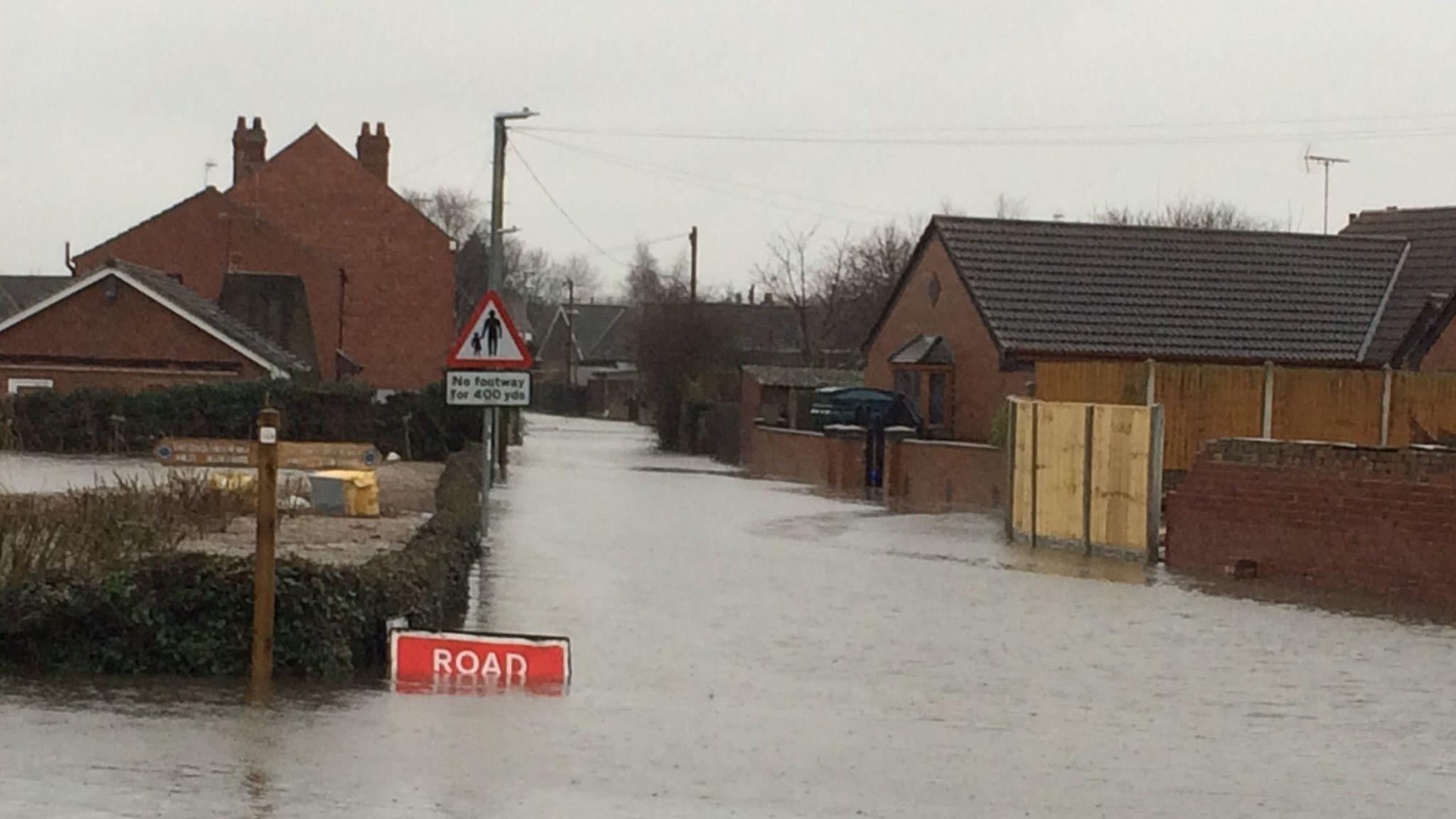 Road covered in flood water. There is a  partly submerged road closure sign. There are properties on the left and right of the road