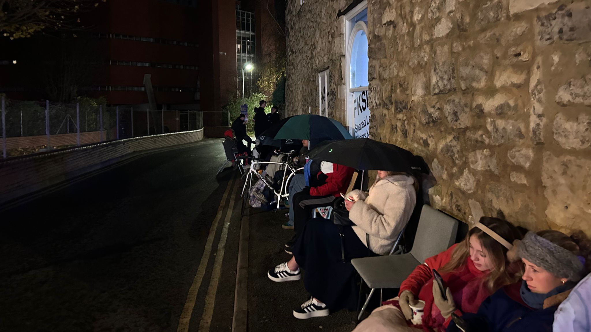 A row of people sitting on chairs along a pavement with the wall of the letting agent building behind them. A lot of the people are wearing coats and hats, and some have umbrellas up.