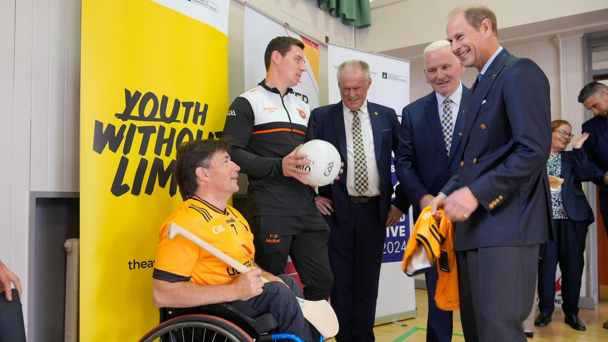 The Duke of Edinburgh, Prince Edward, in a school hall chatting to members of a local GAA team. The Duke has been presented with a GAA jersey. There is a yellow sign behind them that says 'Youth Without Limits' in black writing.