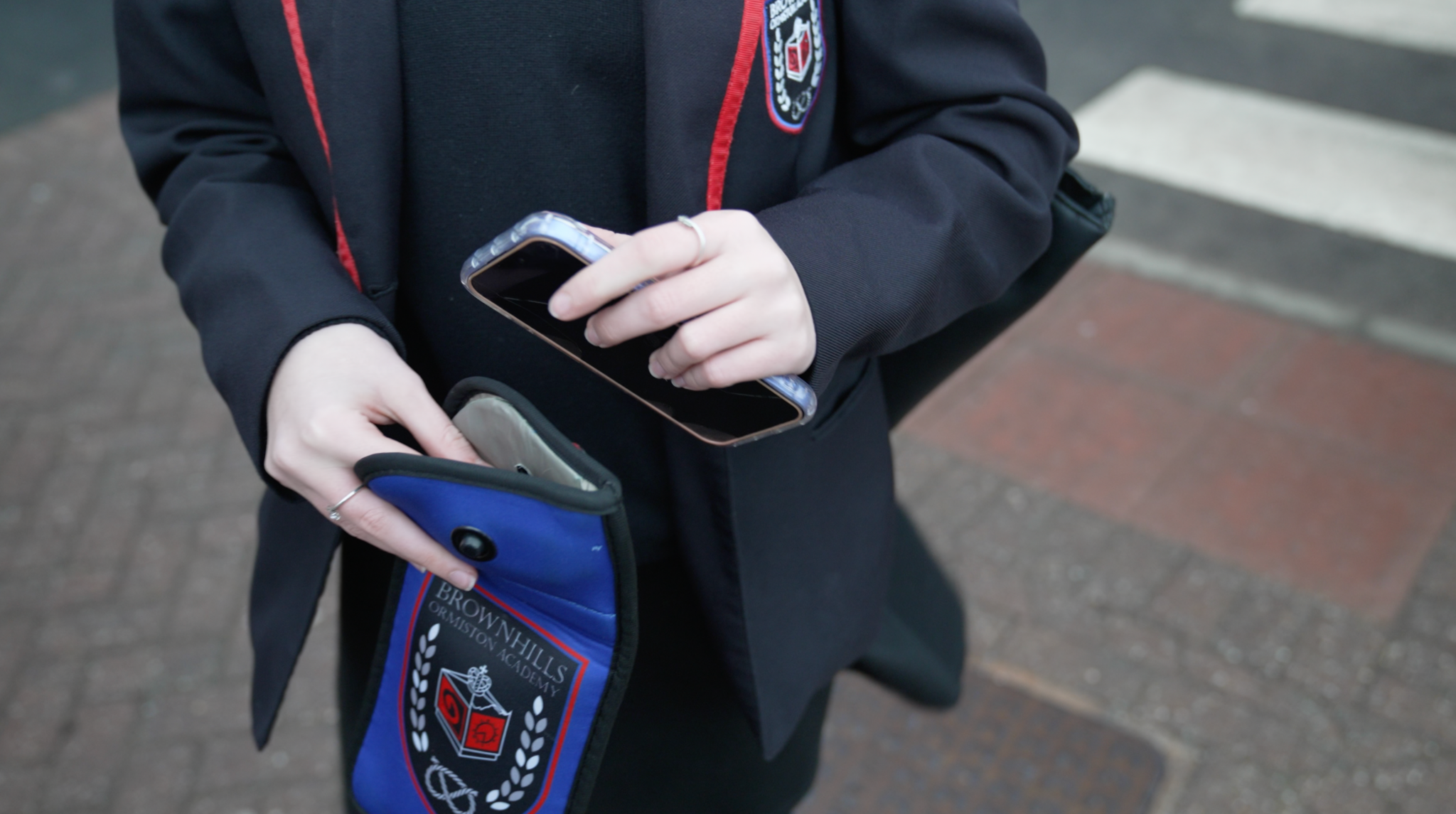 Close-up of a pupil successful  a acheronian  schoolhouse  blazer with reddish  trim placing a smartphone into a tiny  bluish  pouch bearing the schoolhouse  crest, lasting  connected  a pavement.