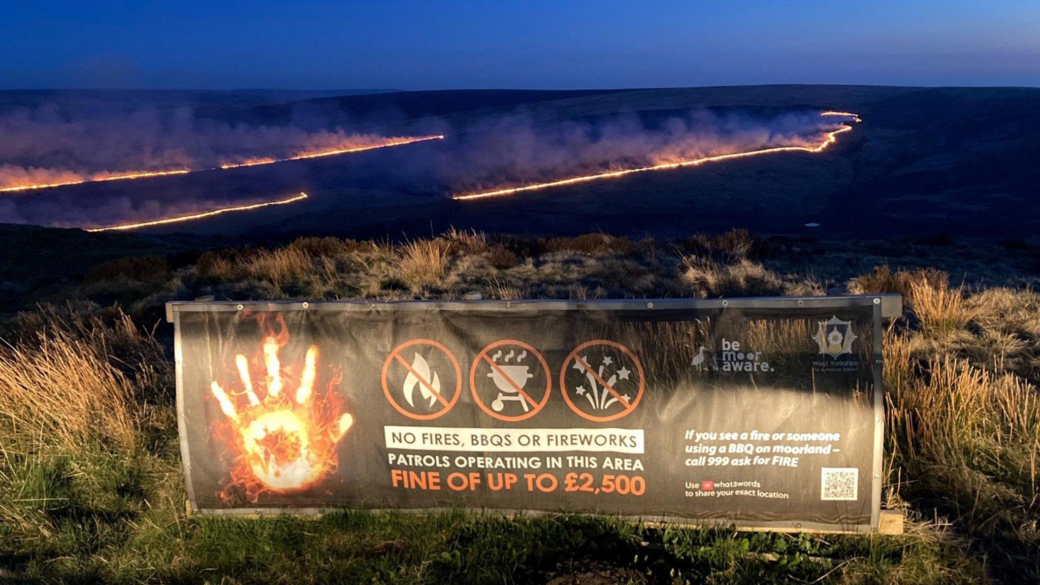 A large wildfire fire in a giant ring shape, with smoke drifting into the sky, next to a sign warning about BBQs, fireworks and camp fires.