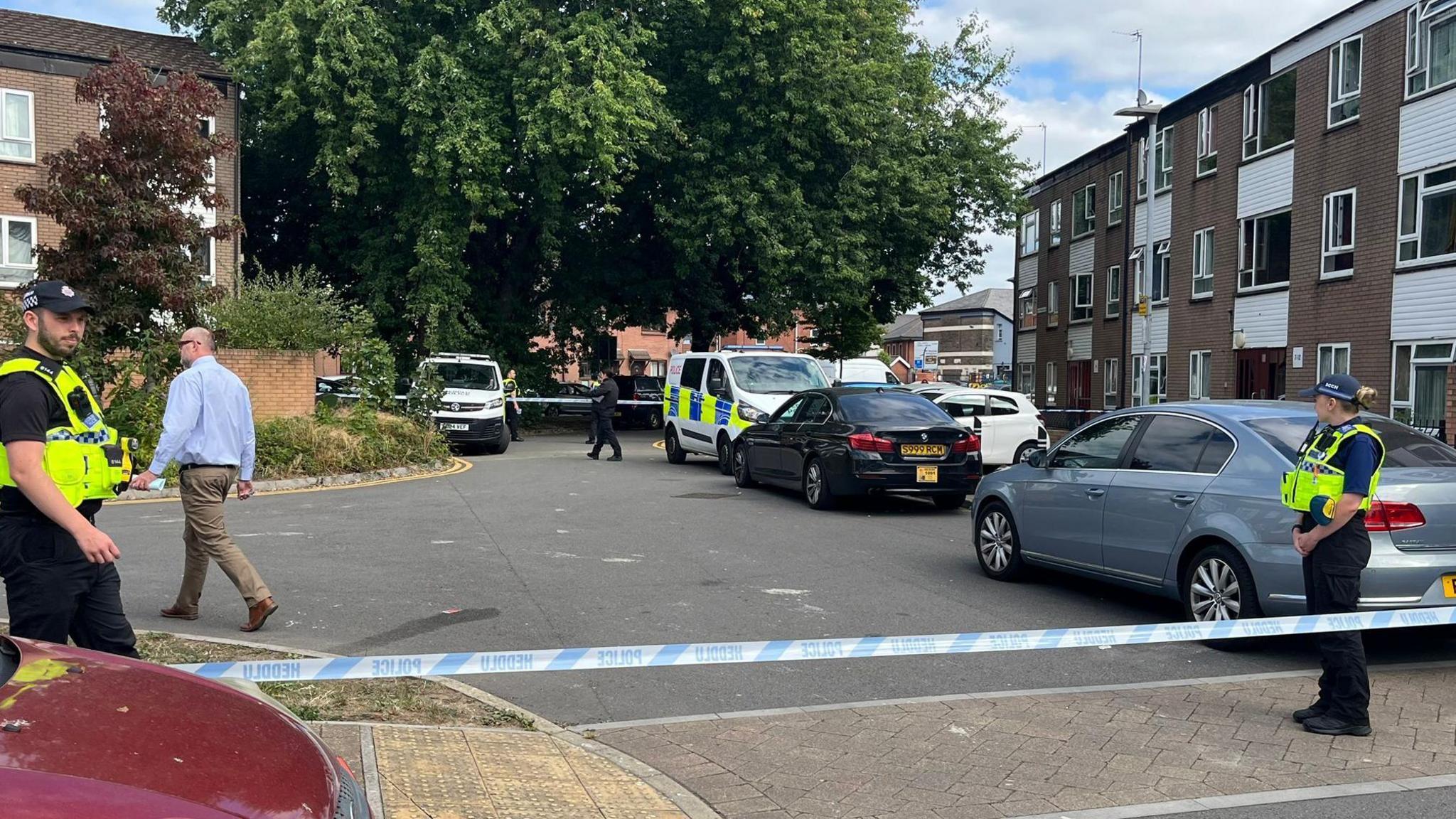 Low rise block of flats with cars parked outside and police van. Two police officers in uniform stand either side of police tape cordoning off the area