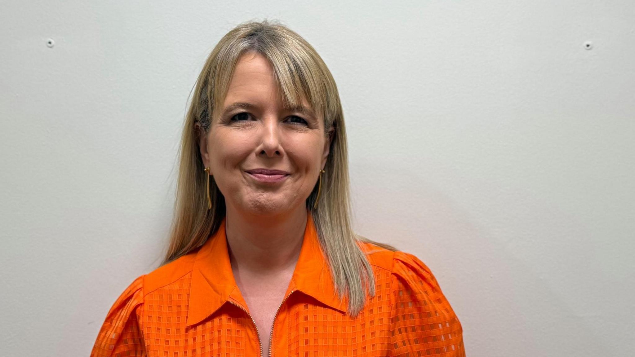 Tracey Bleakley looks direcly at the camera. She has blonde mousey hair with a side fringe. Her hair is just past her shoulders and is worn down. She has a bright orange collared blouse on and is smiling.