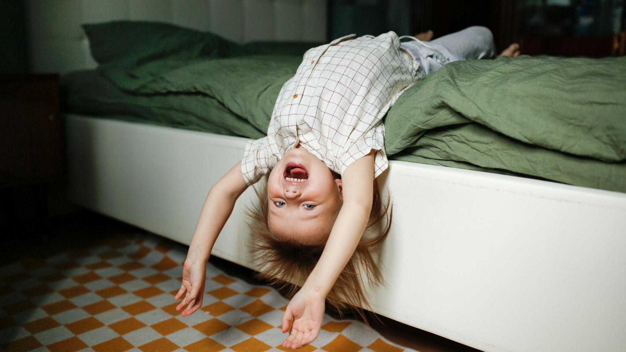 A young child smiling, while hanging upside down from the bed
