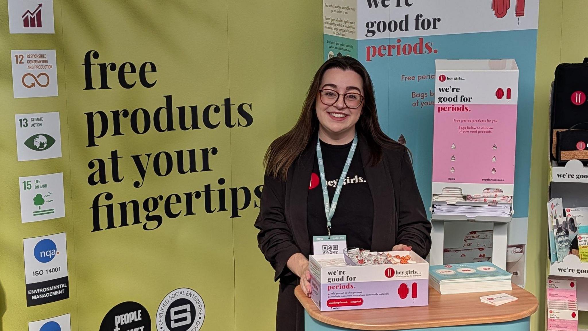 woman with brown hair and glasses smiling at a stand. Behind here there are period products