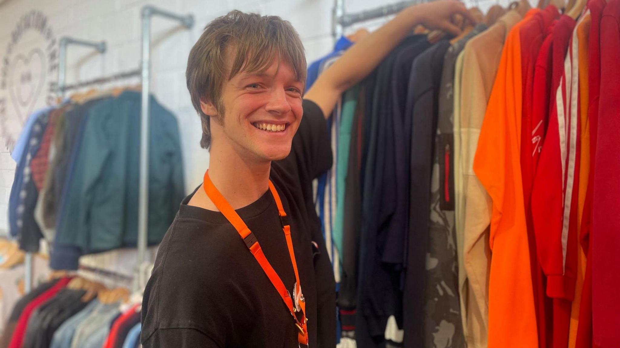 A smiley young man with floppy hair flicks through a rail of brightly coloured vintage clothing