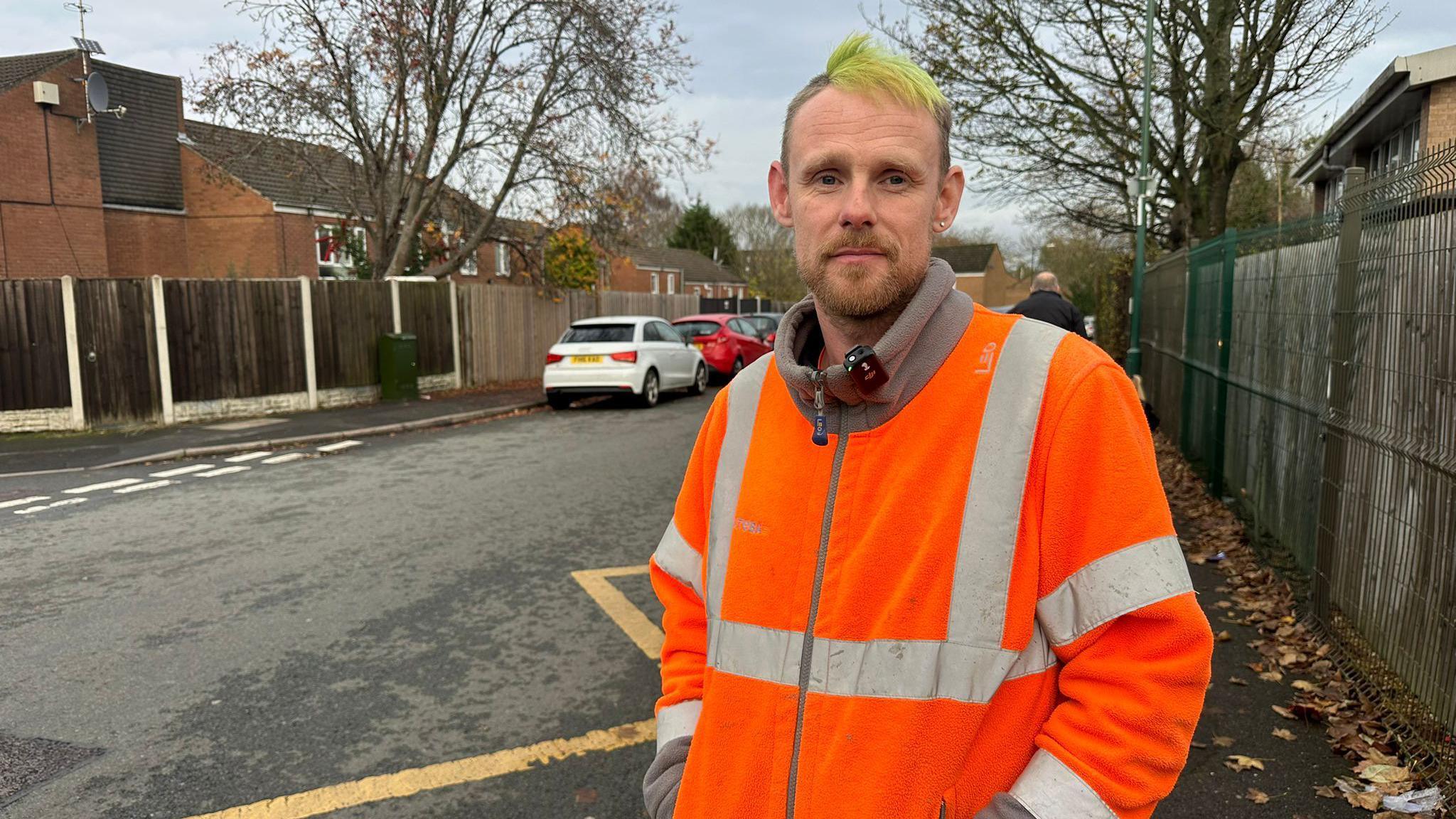 A man wearing a high vis jacket. He has green hair. He is stood in front of a road with cars parked along it.