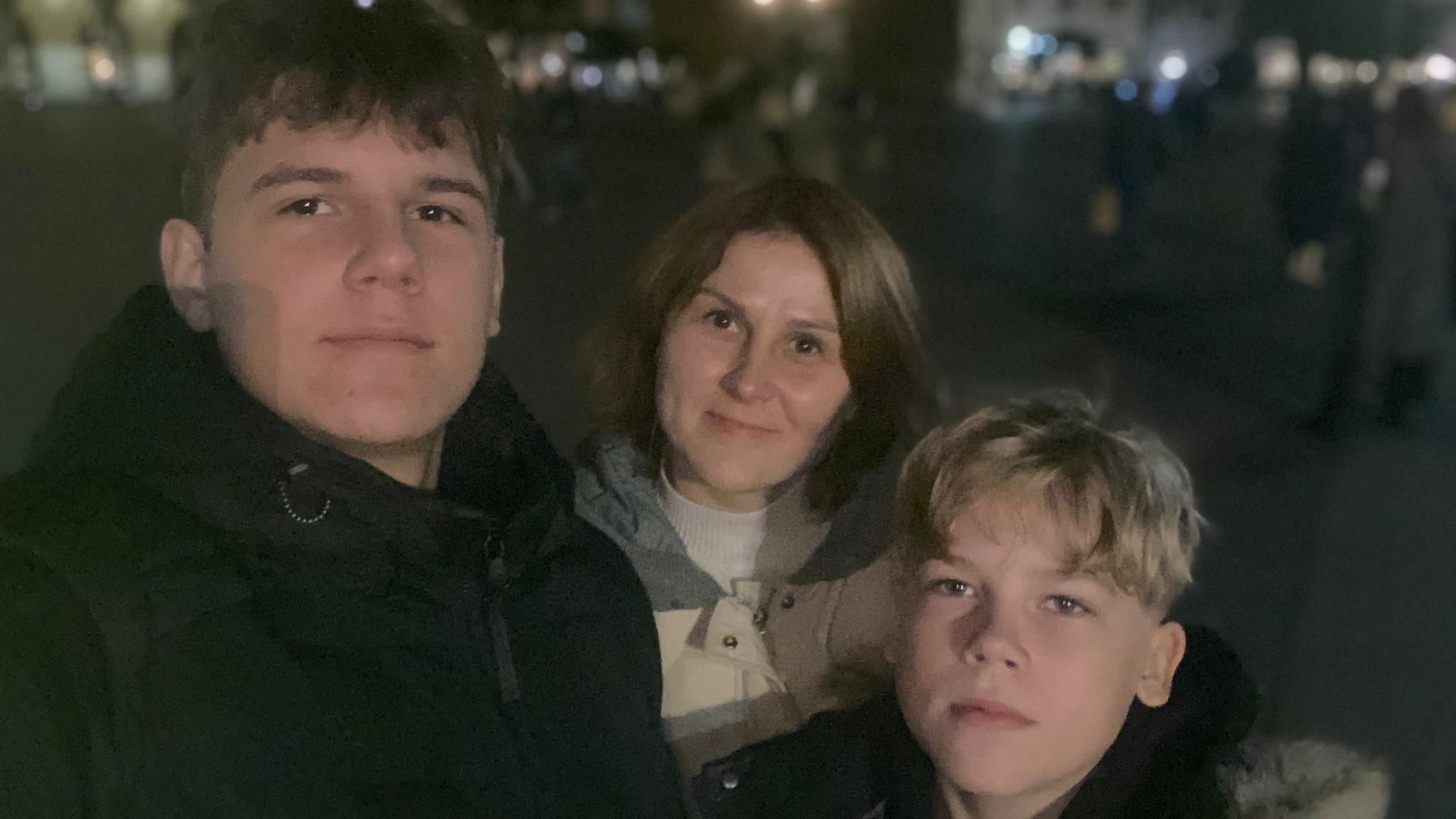 The picture shows a woman, Iryna Maracine, pictured inbetween her 12 and 16 year old sons. It is night time, they are outside and wear winter coats.