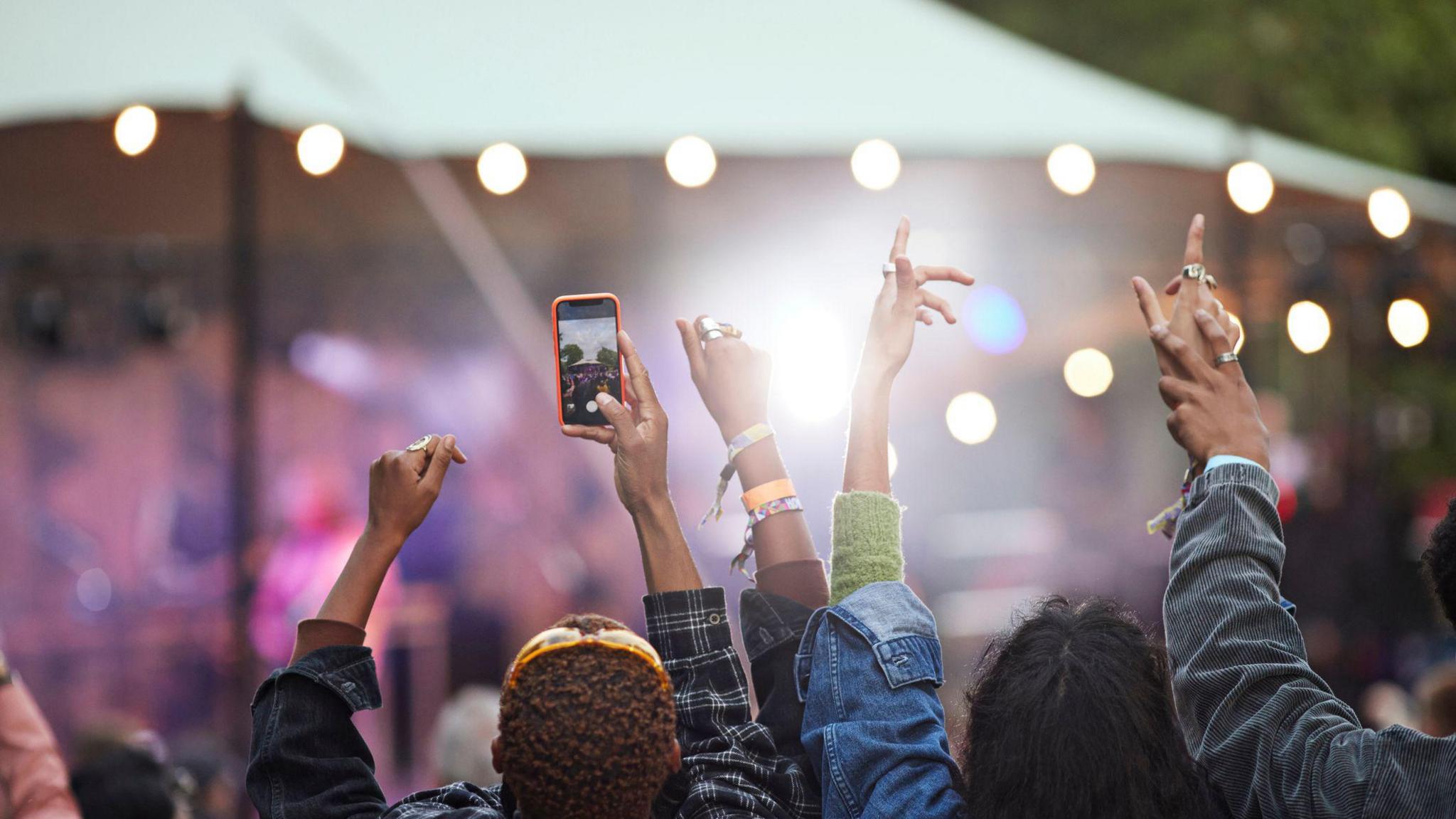A rear view of a crowd at a music festival. People have their arms up celebrating while one is recording on their phone.
