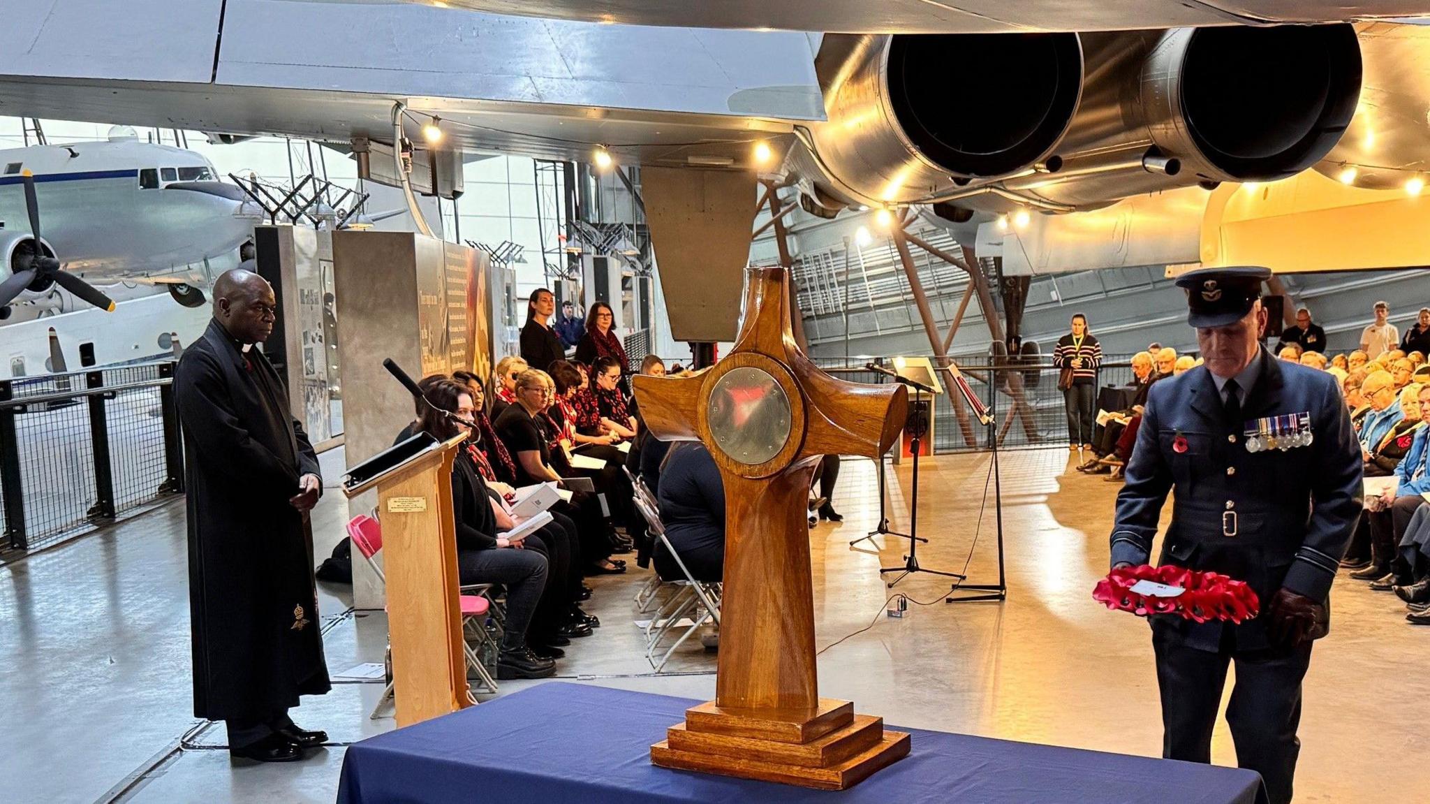 A man in blue military uniform with his head bowed and carrying a poppy wreath approaching a large wooden cross on a blue table with other people in dark clothing behind him